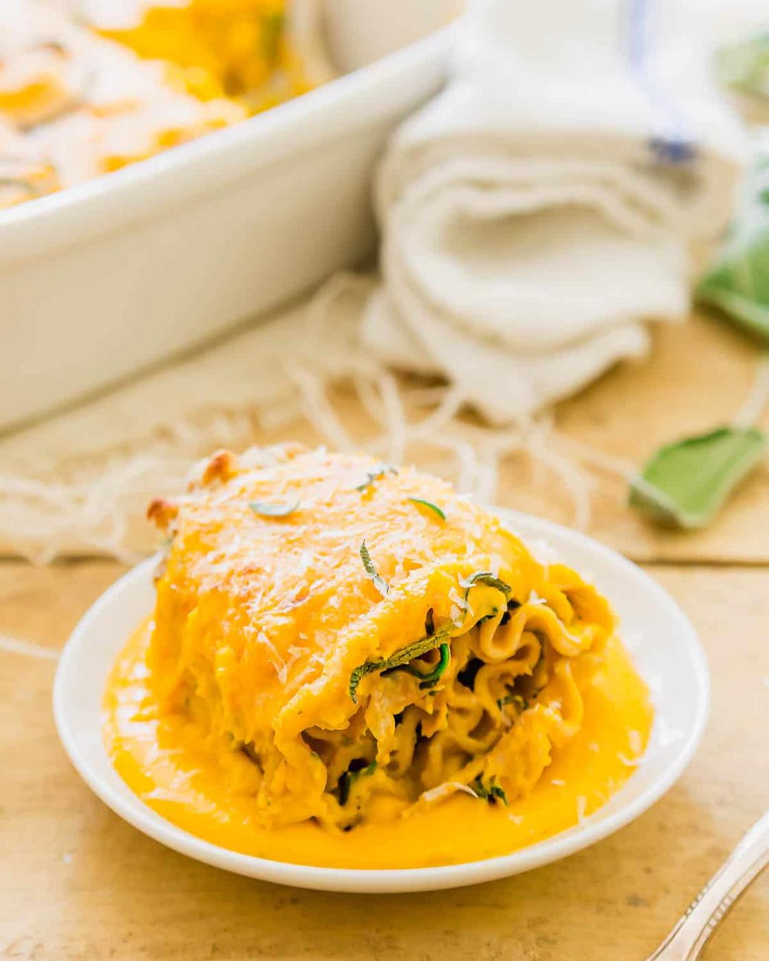 A plate with a lasagna rollup topped with creamy butternut squash sauce and fresh herbs. The lasagna is surrounded by a thick, rich sauce. In the background, a white cloth napkin and casserole dish are slightly visible on a wooden surface.