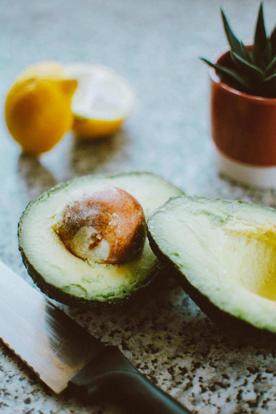 Sliced avocado and lemons on a kitchen countertop with a knife and potted plant in the background.