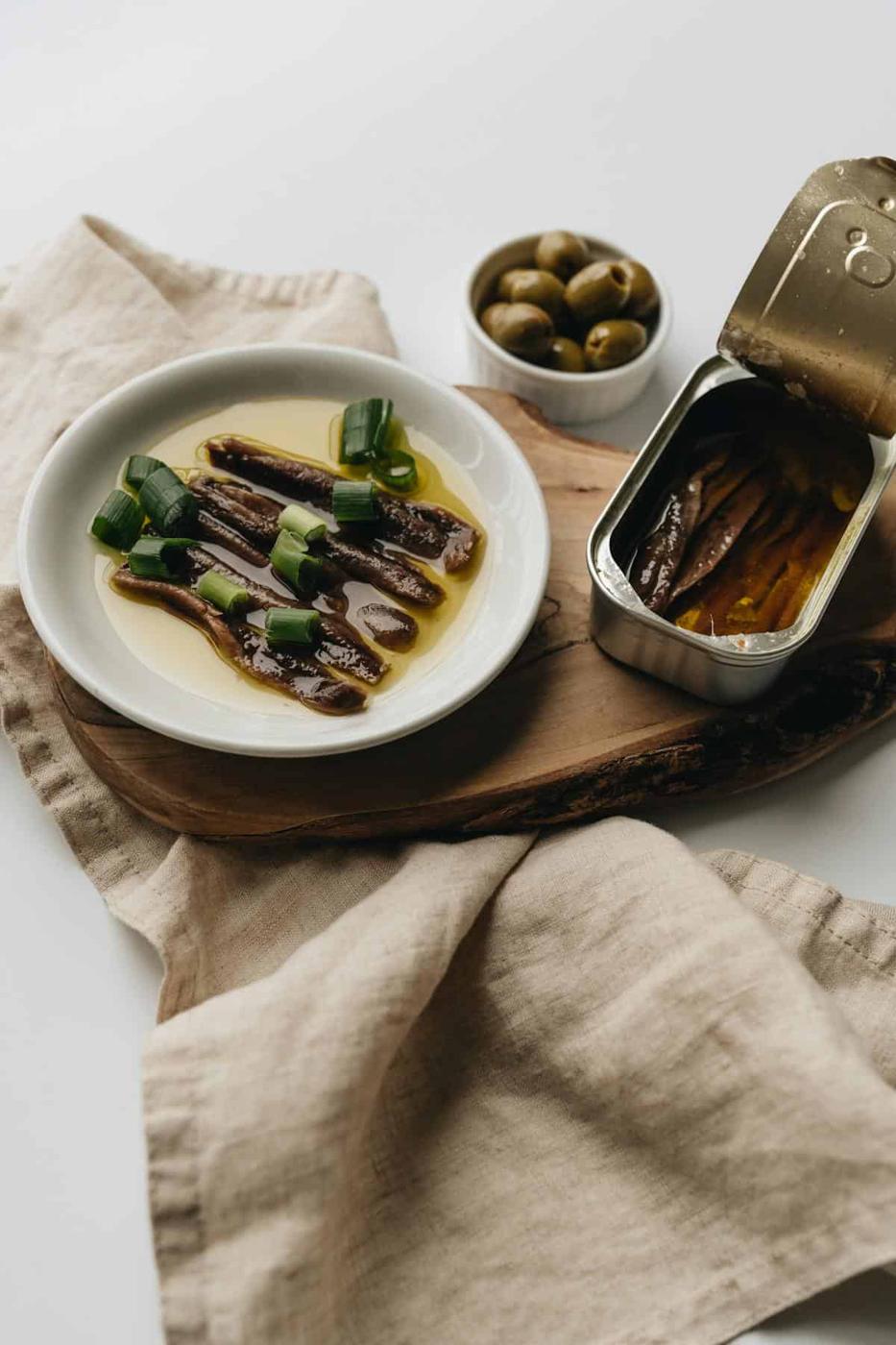 A white plate with anchovies in oil, garnished with green onions, sits on a wooden board beside an open tin of anchovies and a small bowl of green olives. A beige cloth napkin is underneath.