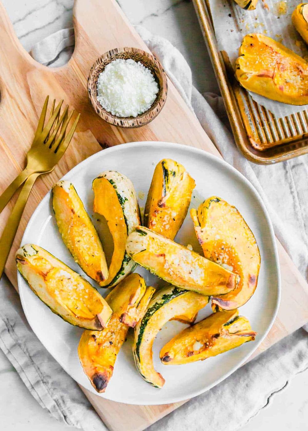 Overhead shot of roasted parmesan carnival squash on a white plate with gold forks on the side and small bowl of grated cheese.
