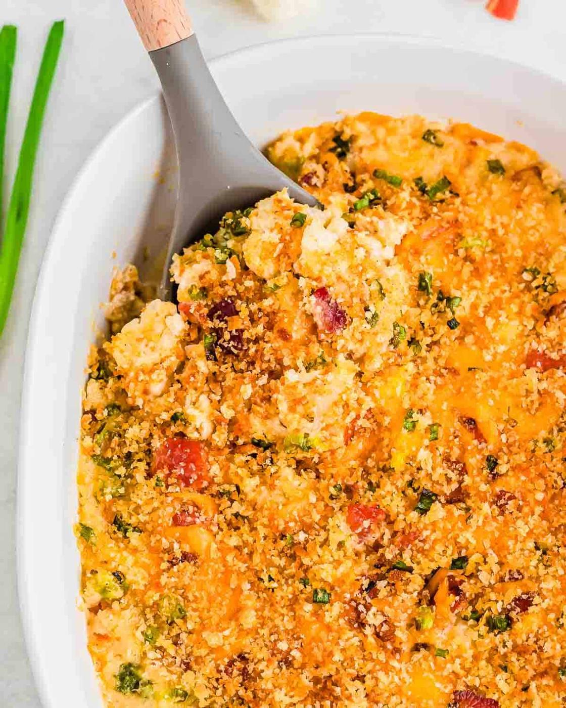 A close-up of a gratin with a golden breadcrumb topping, visible vegetables, and melted cheese in a white oval dish. A grey spoon is scooping out a portion. Green onion stalks are in the background.