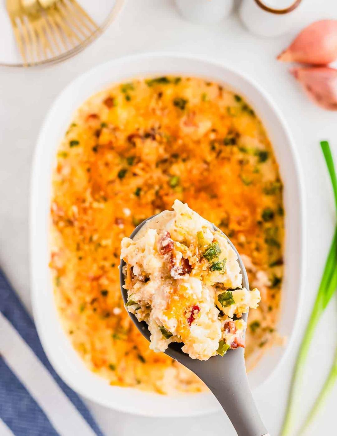A close-up of a spoon lifting a serving of creamy baked cauliflower gratin with visible chunks, green herbs, and crispy topping, held over a white baking dish. Ingredients like shallots and green onions are on the side.