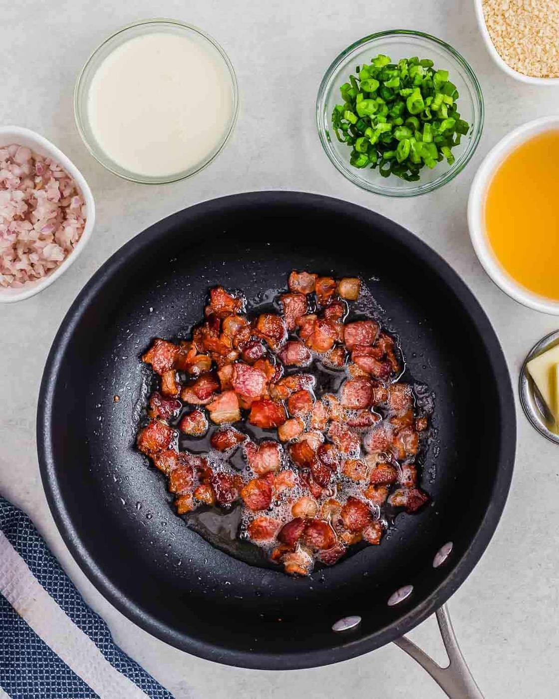 A frying pan with crispy bacon pieces in the center. Surrounding the pan are small bowls containing sliced green onions, cream, chopped shallots, broth, and a block of butter. A striped cloth is partially visible to the side.