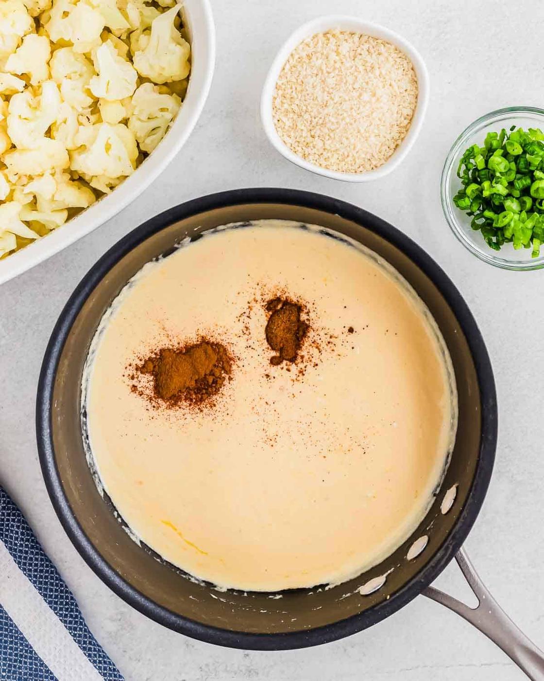 A saucepan filled with creamy cheesy sauce, topped with two piles of spices, likely paprika or chili. Nearby are bowls of chopped green onions, breadcrumbs, and a dish of cooked cauliflower. A striped cloth sits on the left side.