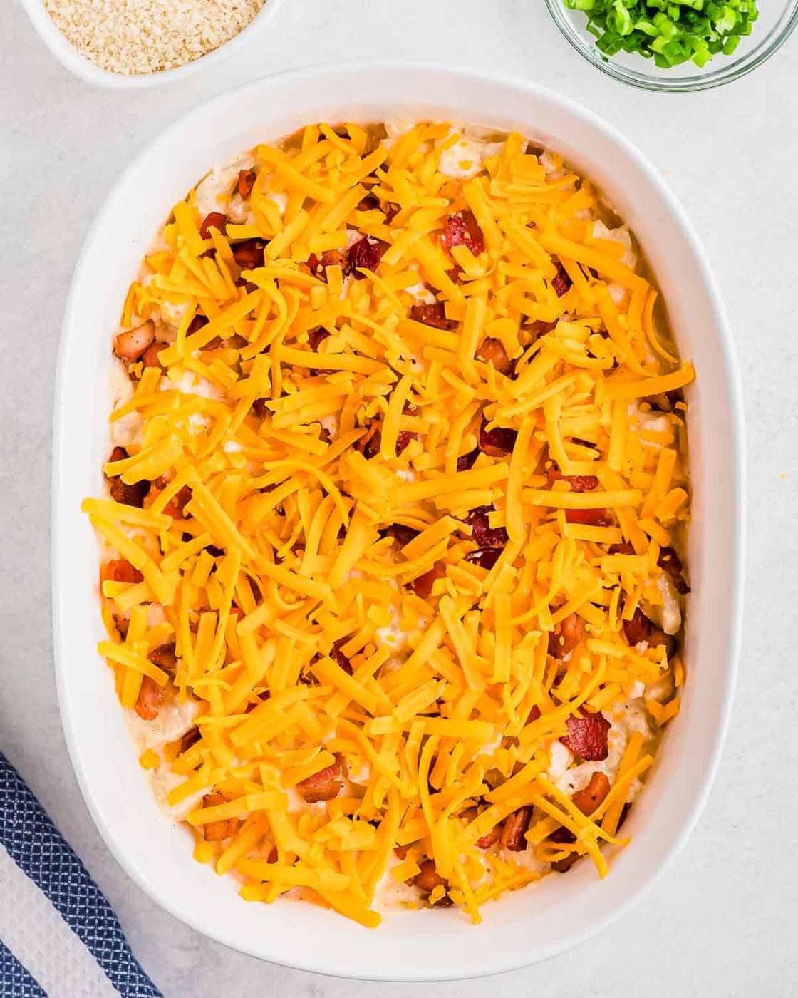 A white oval baking dish filled with a creamy mixture of cauliflower, topped with shredded cheddar cheese and bits of bacon, set on a light-colored countertop beside a blue-striped cloth.