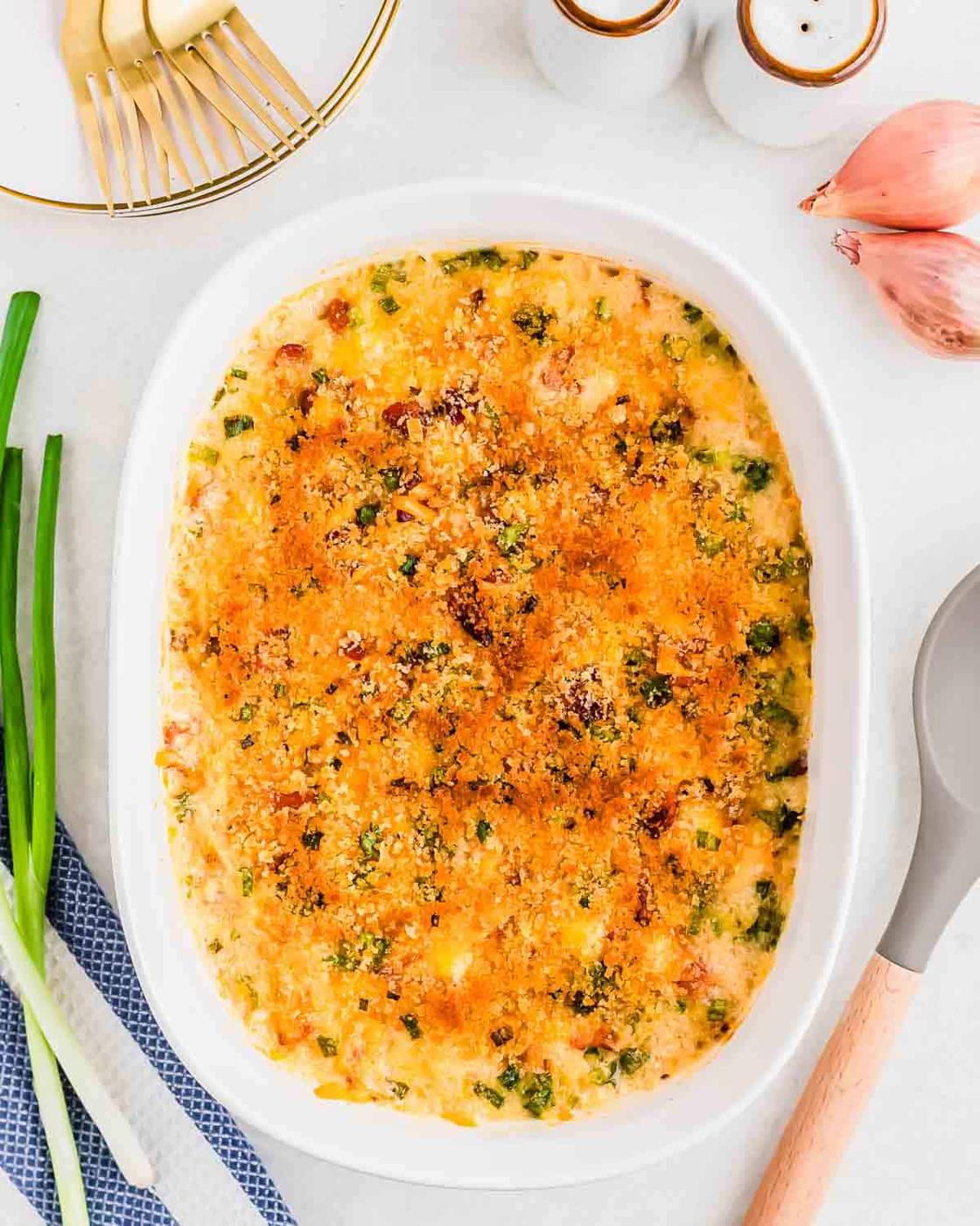 Baked cauliflower gratin with a golden breadcrumb topping in a white oval dish. Surrounding it are green onions, a shallot, and a spoon. A fork with a lemon slice and salt and pepper shakers are nearby on a light surface.