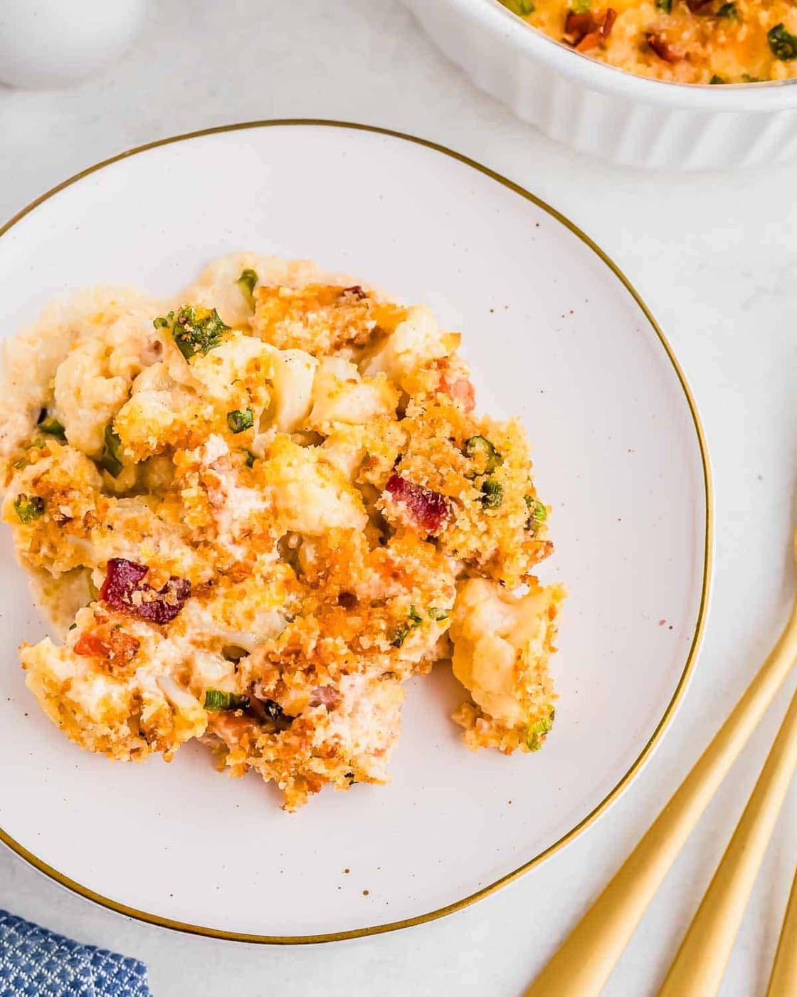 A serving of cauliflower bacon gratin topped with crispy, golden breadcrumbs and bits of bacon on a white plate with a gold rim. Forks are to the side.