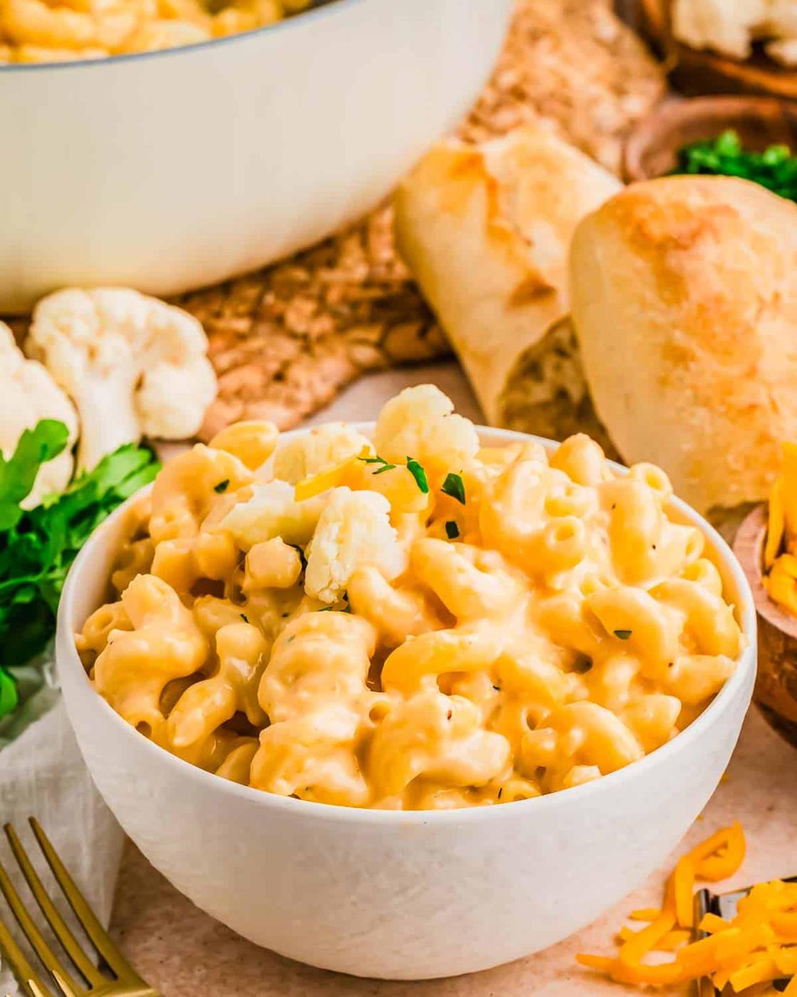 A white bowl filled with creamy macaroni and cheese, topped with cauliflower florets and parsley, sits on a table next to bread, shredded cheese, and fresh herbs.