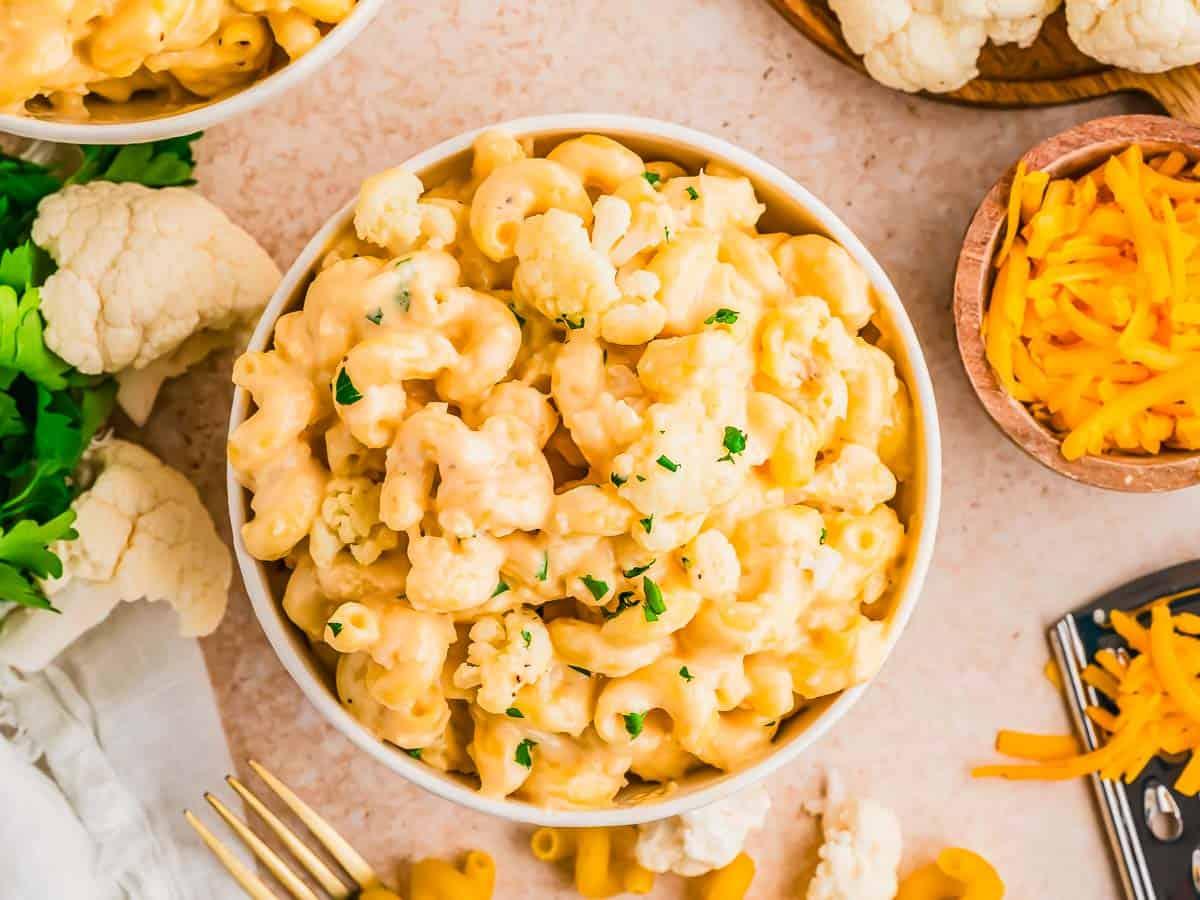 A bowl of creamy mac and cheese garnished with chopped herbs sits on a table, surrounded by shredded cheddar cheese, cauliflower florets, parsley, and a fork.