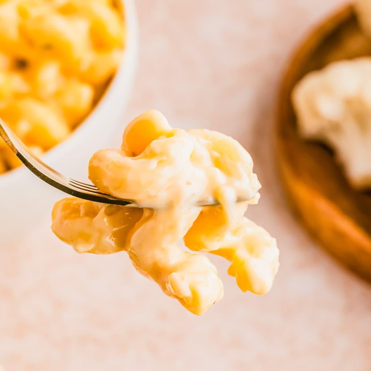 A close-up of a fork holding a creamy bite of cauliflower mac and cheese, with a bowl of mac and cheese in the background on a light surface.