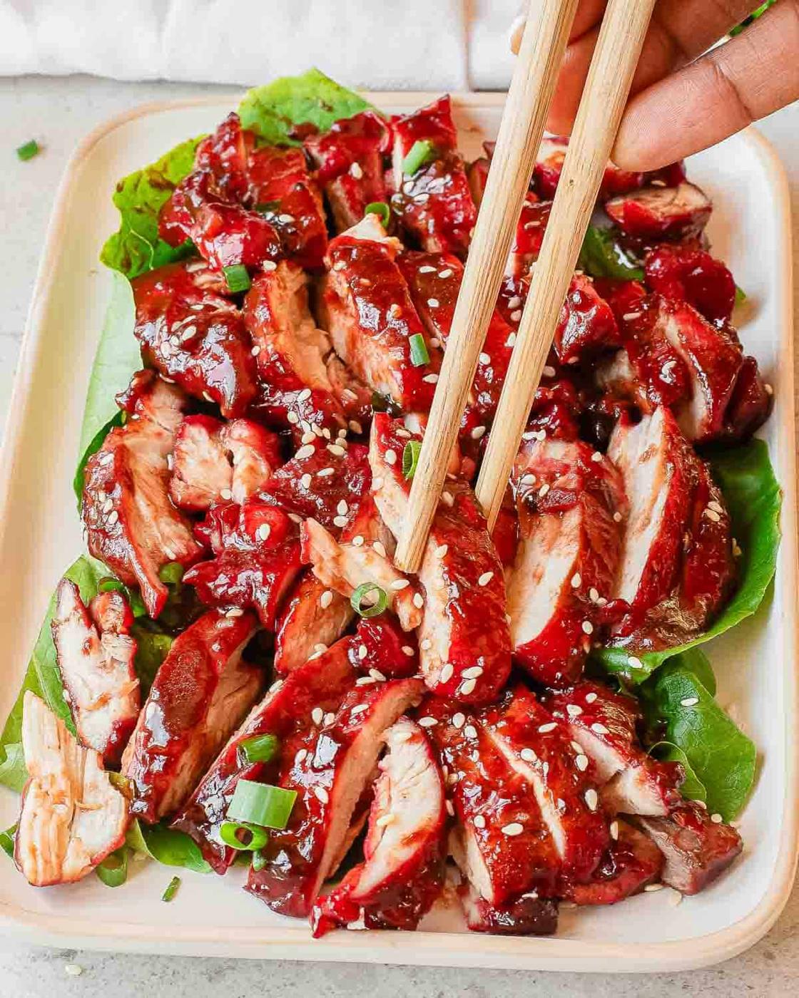 A plate of sliced, char siu glazed baked chicken topped with sesame seeds and chopped green onions, served on a bed of leafy greens. A hand is picking up a piece of chicken with chopsticks.