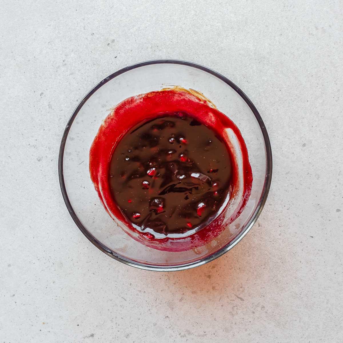 A glass bowl containing a dark brown and red marinade for char siu chicken, sits on a light-colored surface. Some of the mixture is splattered on the sides of the bowl.