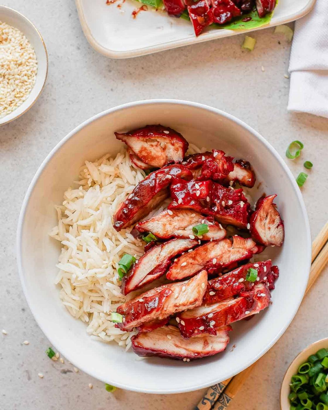 A white bowl filled with steamed white rice topped with sliced chicken char siu, garnished with sesame seeds and chopped green onions. Chopsticks and small dishes of sesame seeds and sauce are nearby.
