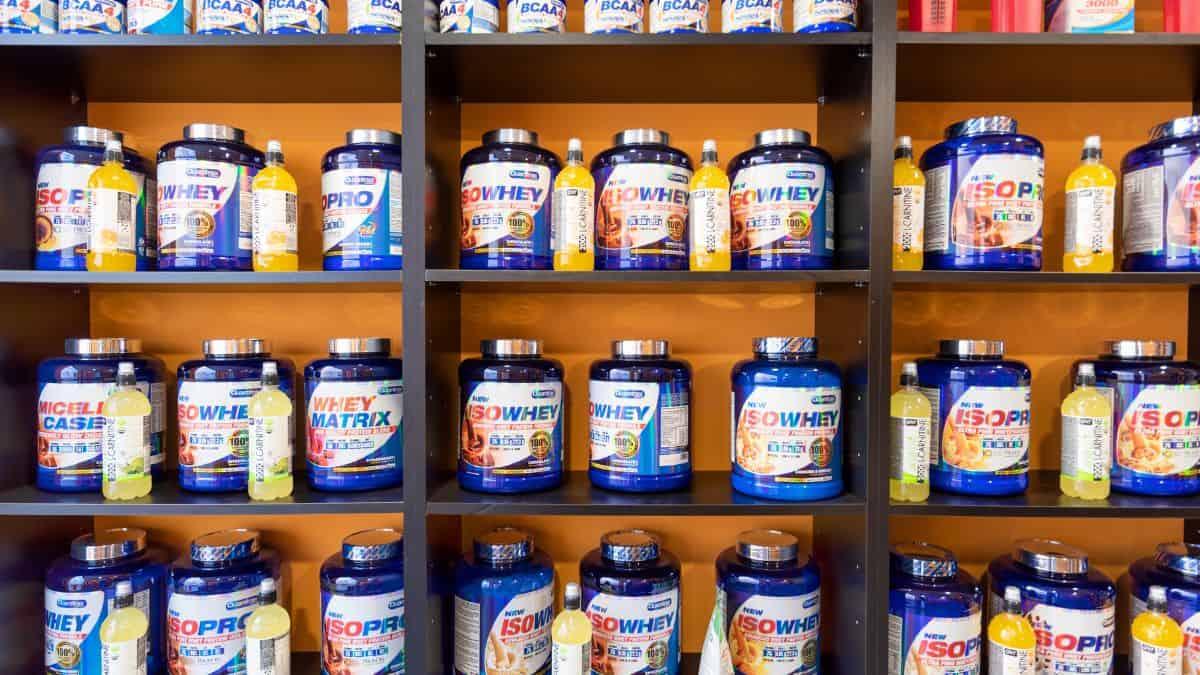 A display of various blue protein supplement containers and bottles of beverages arranged on a black shelf against an orange wall.