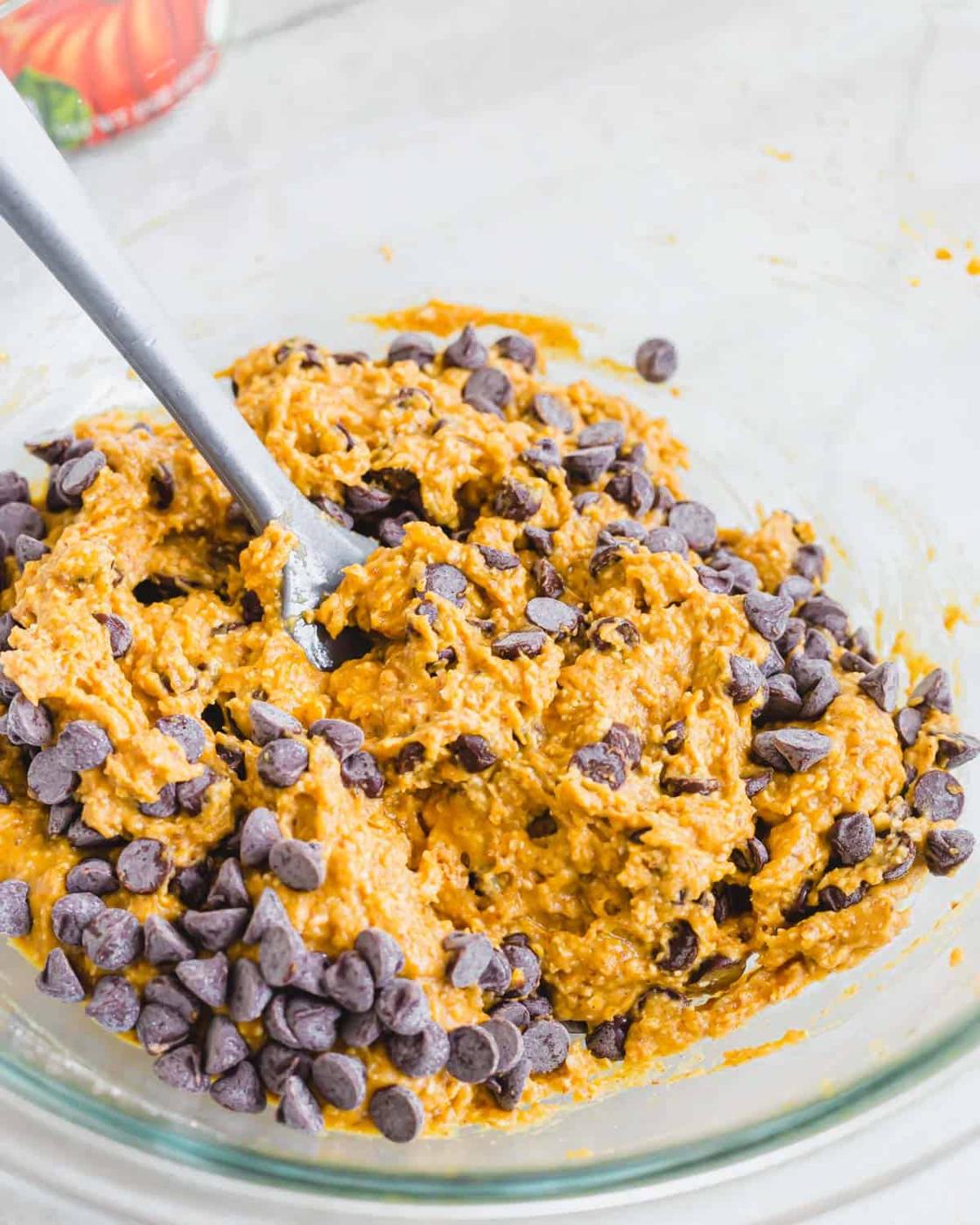 Vegan pumpkin cookie batter in a glass bowl with chocolate chips and a gray spatula.