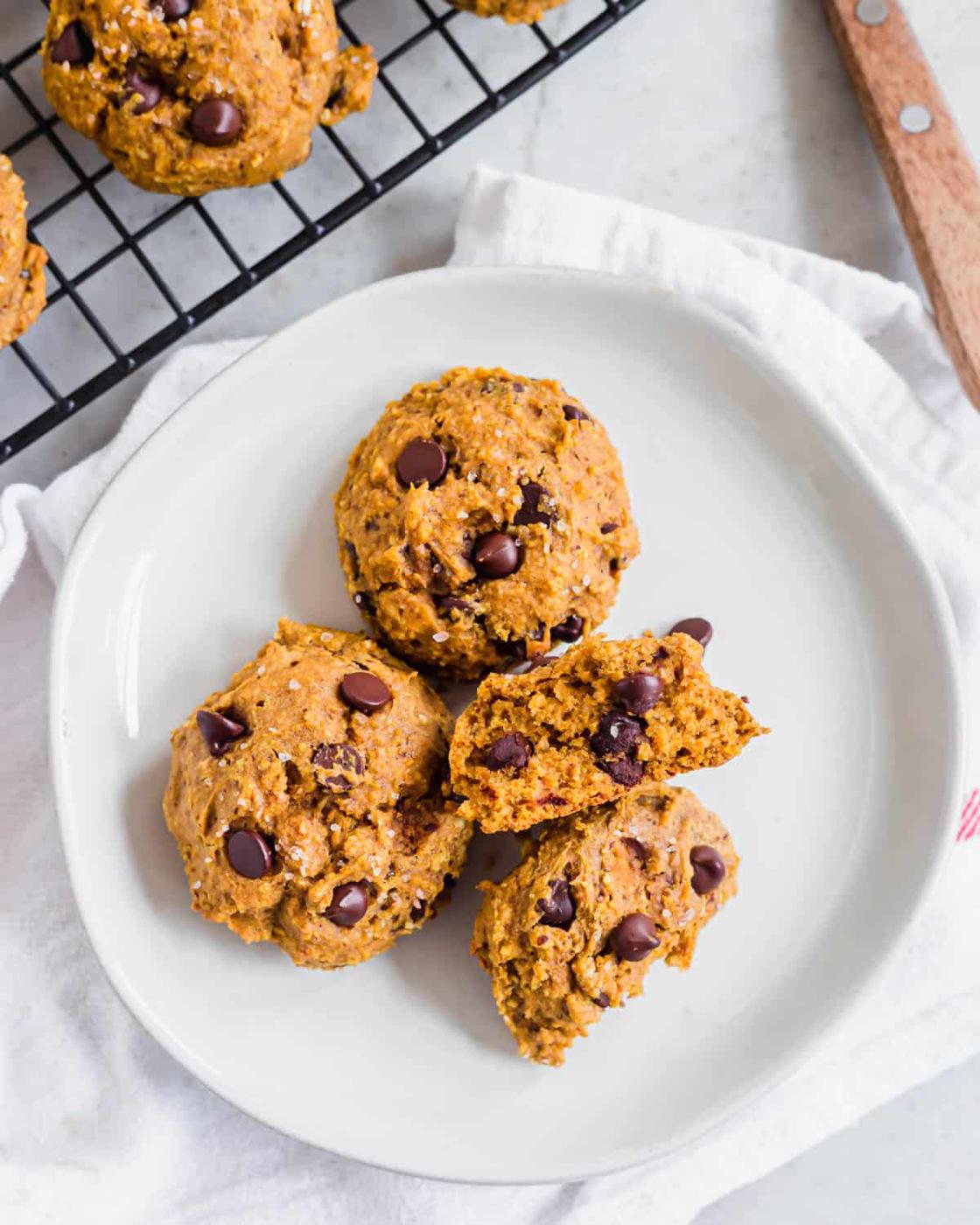 Three chewy pumpkin cookies with chocolate chips on a plate, one broken apart to see the middle.