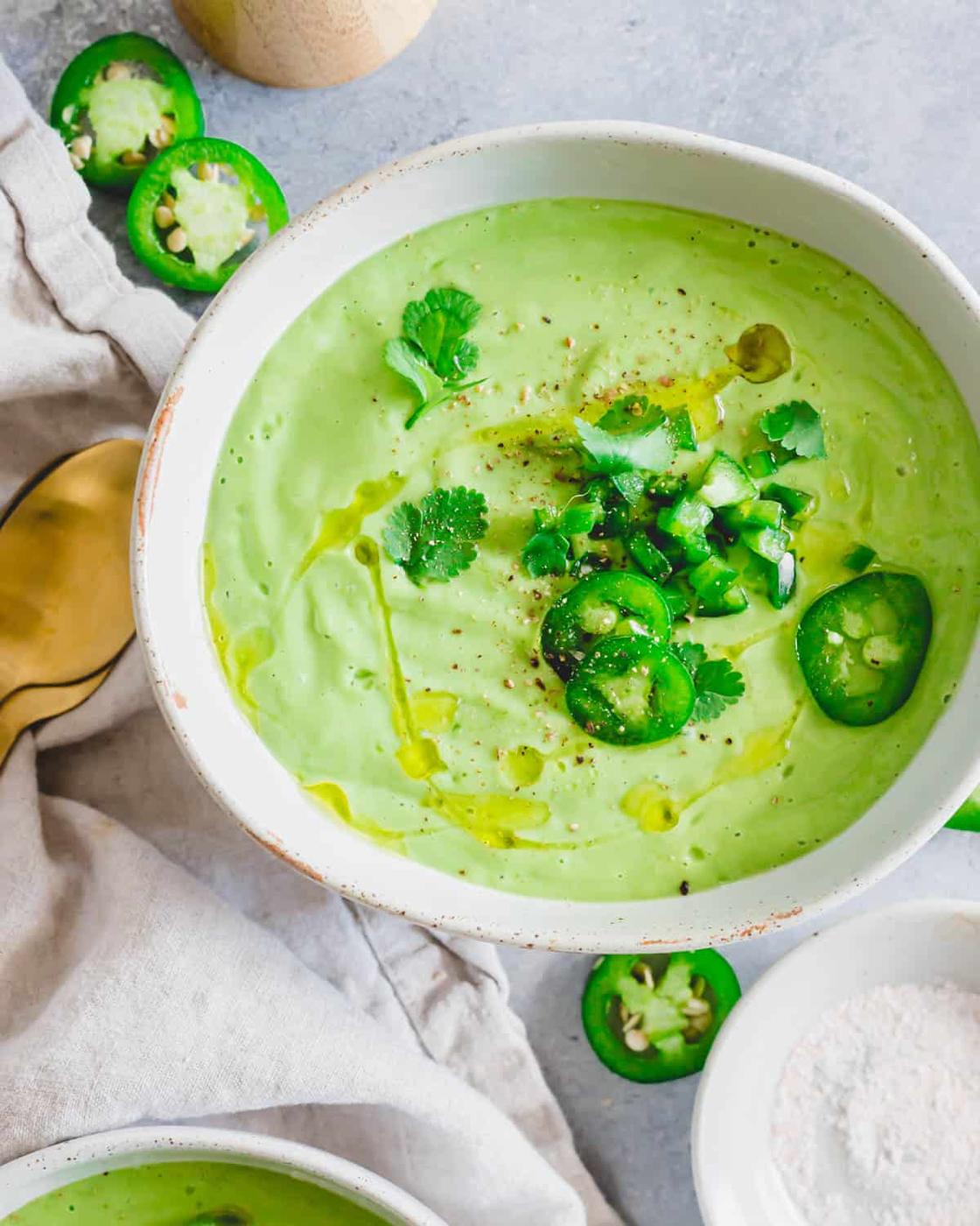 Chilled avocado soup in a bowl drizzled with olive oil and garnished with jalapeño peppers, cilantro, salt and pepper.