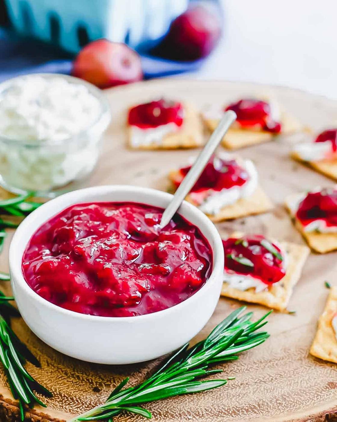 A white bowl of vibrant plum jam with a spoon, placed on a wooden board. Crackers topped with cheese and cranberry sauce are arranged nearby, with fresh rosemary sprigs for garnish. A small glass bowl of white cheese is in the background.