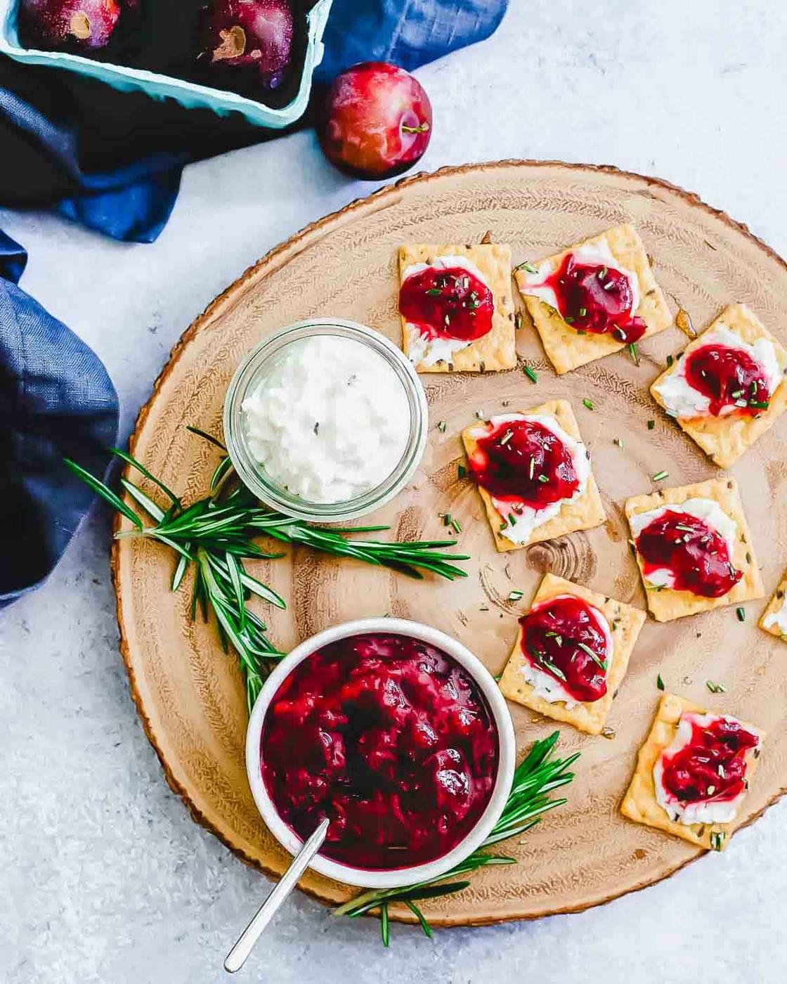 A wooden board with crackers topped with white cheese and plum jam sauce, garnished with rosemary. A jar of white dip and a bowl of berry sauce with a spoon rest beside them. A small dish of plums and a scattered plum are in the background.