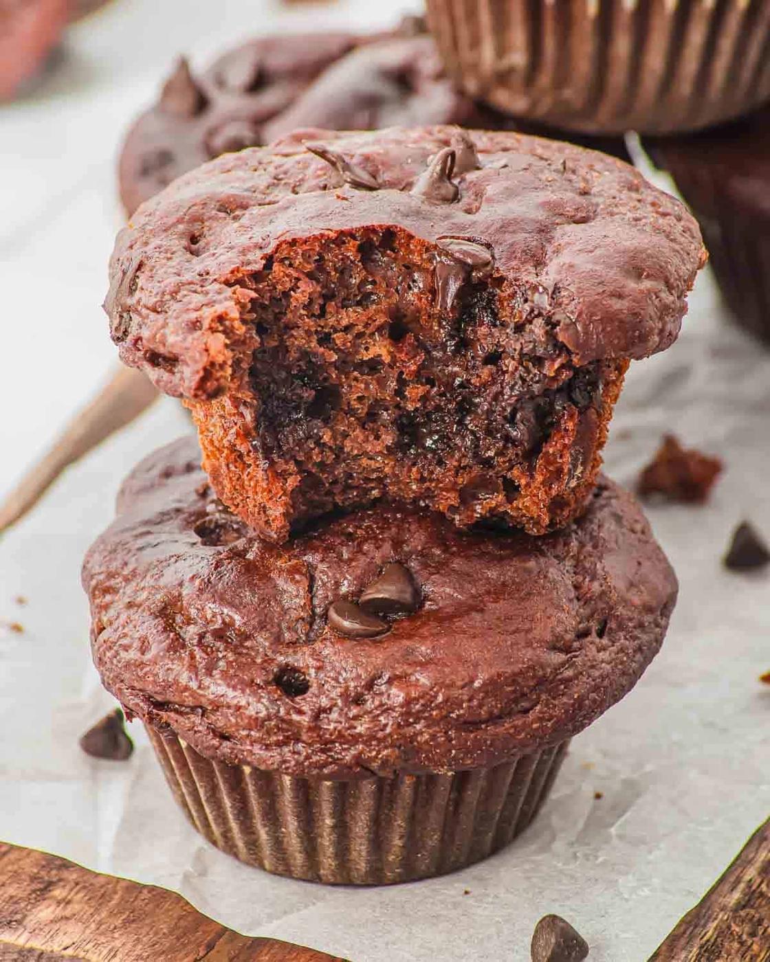 Two chocolate muffins are stacked, with the top muffin showing a bite taken out, revealing a moist, rich chocolate interior. Chocolate chips are scattered on and around the muffins.