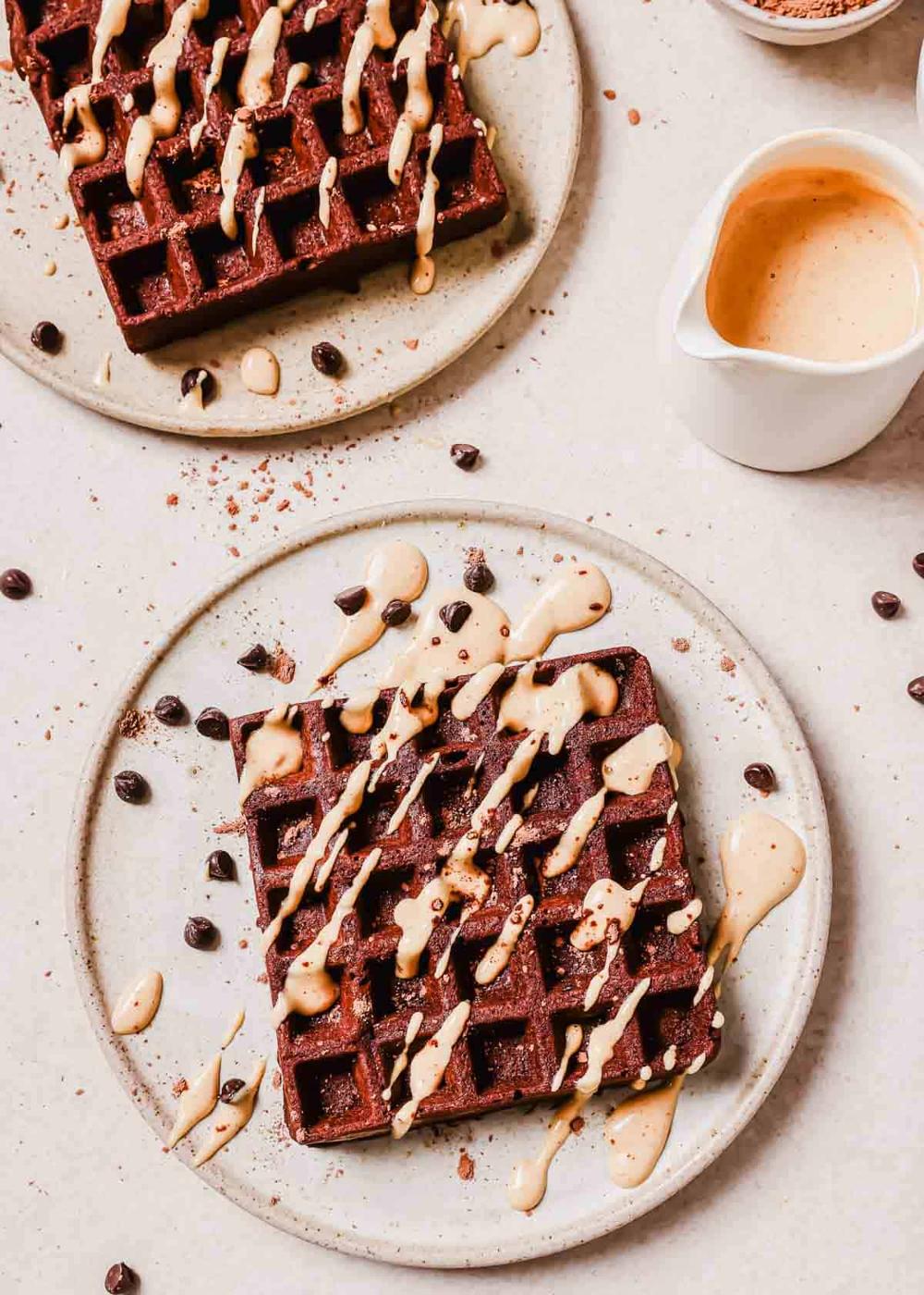 Two plates of chocolate waffles drizzled with peanut butter glaze, garnished with chocolate chips and served with a side of syrup.
