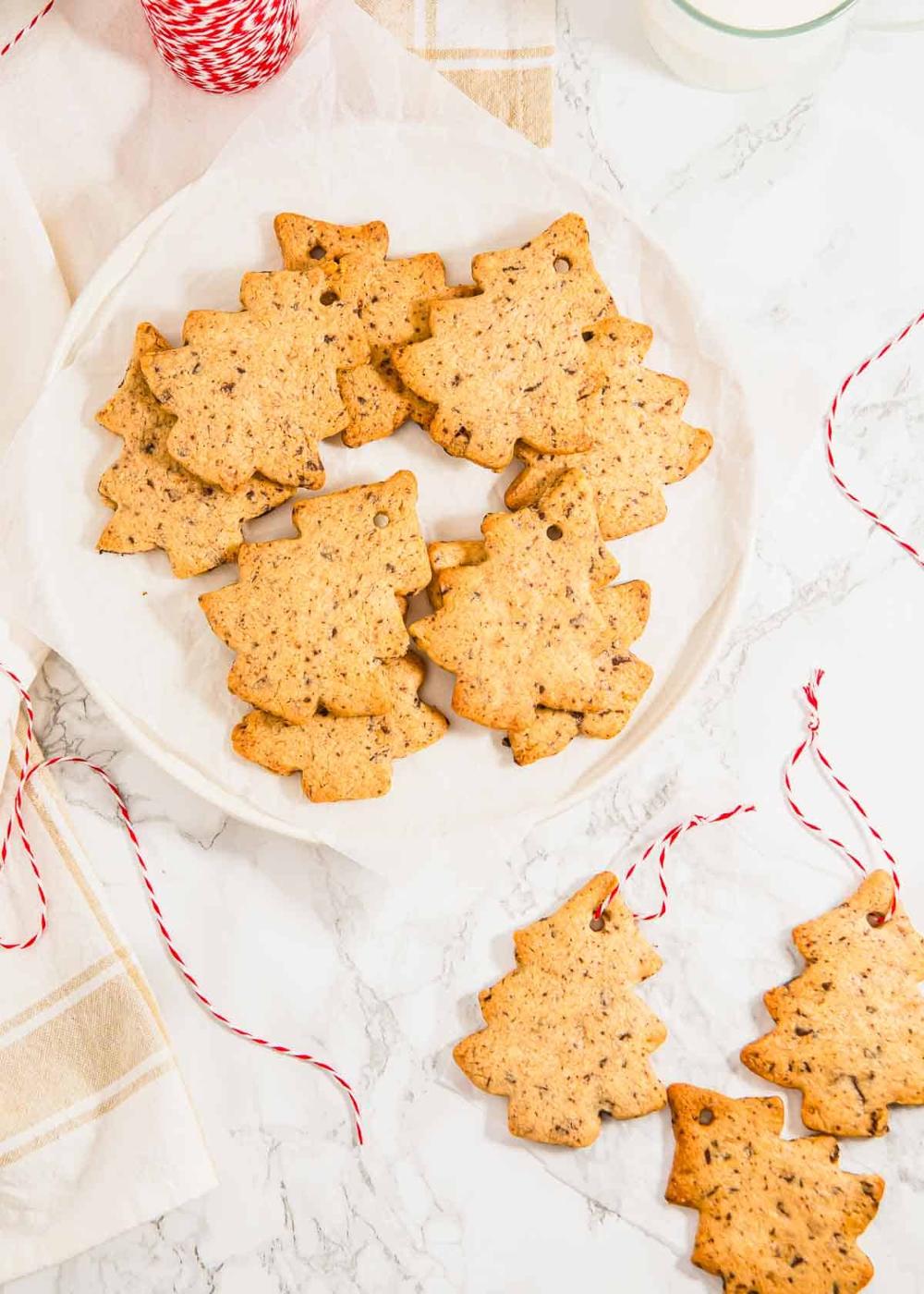 Christmas tree cookies on a white plate with others scattered on the side with red and white string strung through a hole in the top to make them into ornaments.