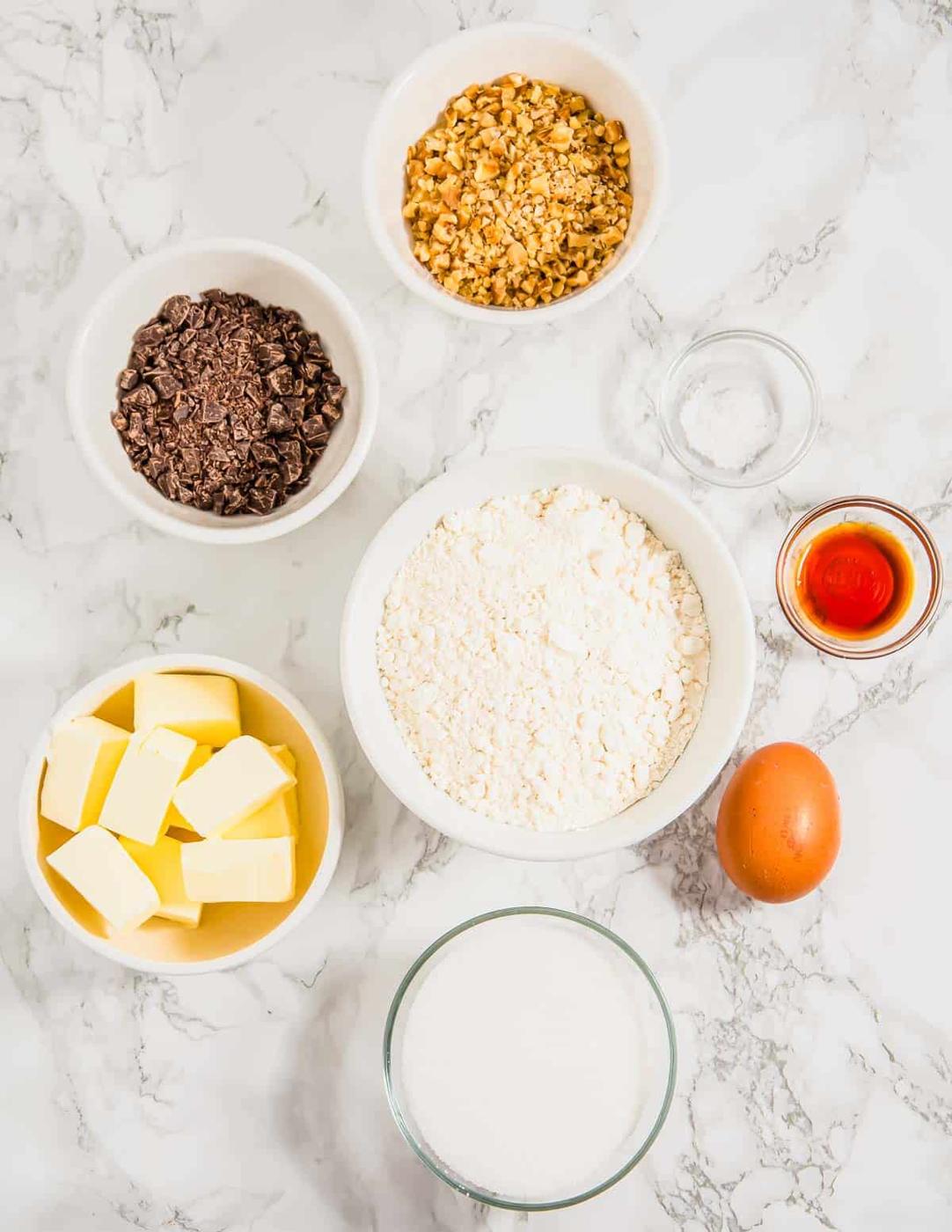 A marble surface with baking ingredients: chopped nuts, chopped chocolate, flour, sugar, butter cubes, an egg, vanilla extract, and salt, each in separate bowls.