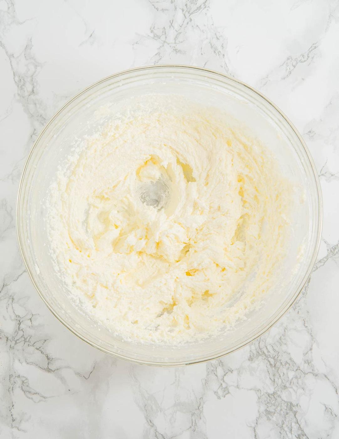 A glass bowl filled with creamed butter and sugar sits on a marble countertop. The mixture has a light, fluffy texture with a smooth surface, indicating it's well-mixed.
