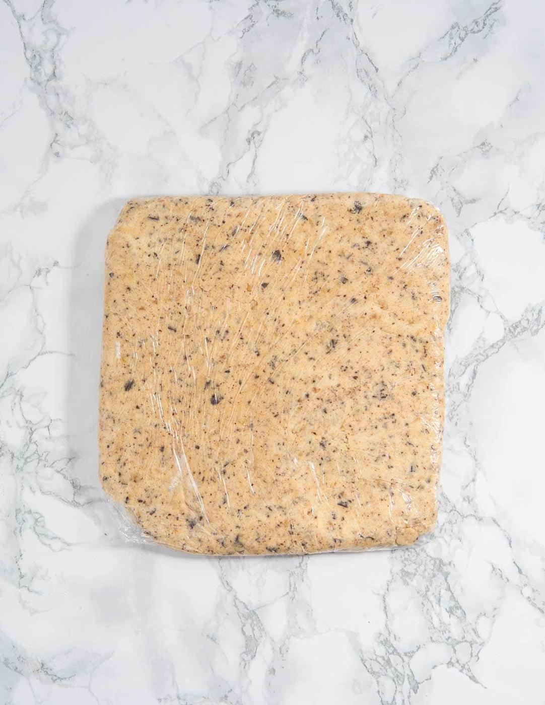 Cookie dough shaped into a rectangle and wrapped in plastic wrap sitting on a marble surface.