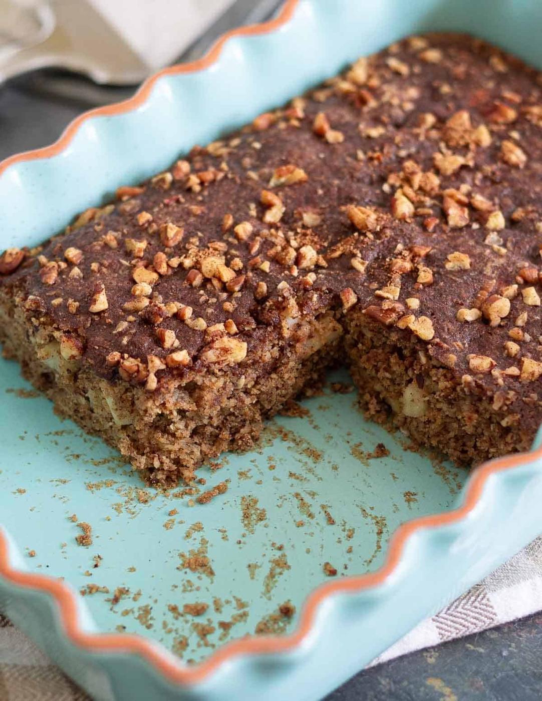 A rectangular baked apple cinnamon cake with a crunchy walnut topping is shown in a light blue ceramic dish. A large piece has been cut out, revealing the moist, crumbly texture inside. Crumbs and chopped nuts are scattered around the dish.