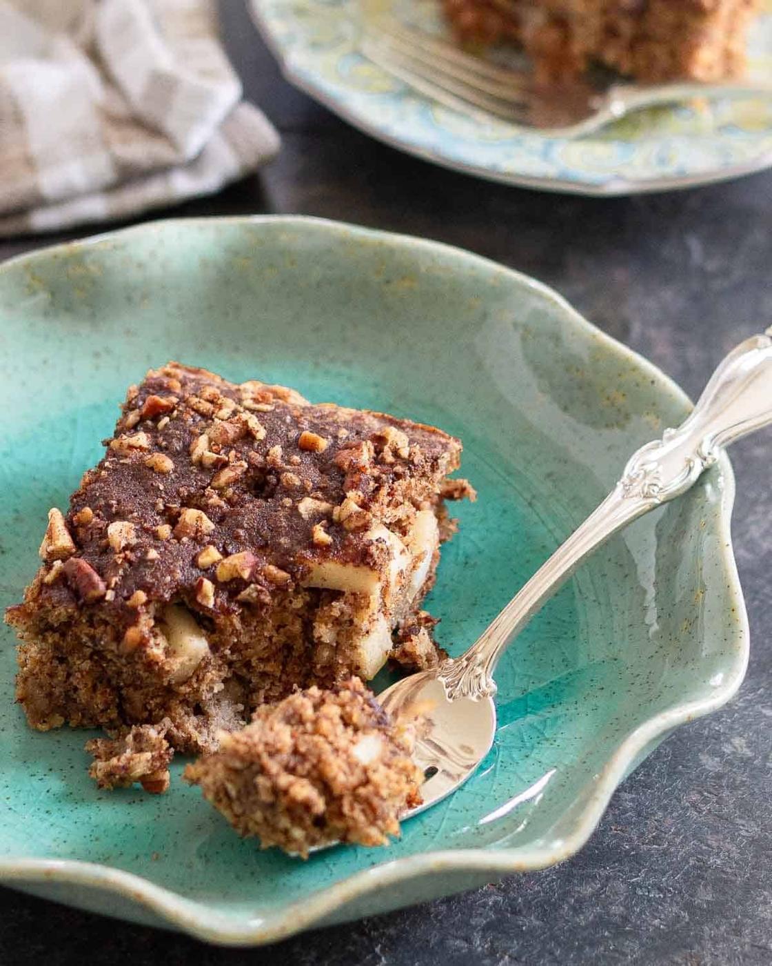 A slice of crumbly cinnamon apple cake with chopped nuts and a cinnamon topping sits on a blue-green plate with a silver spoon, with a forkful cut and ready to eat. Another serving is visible in the background.