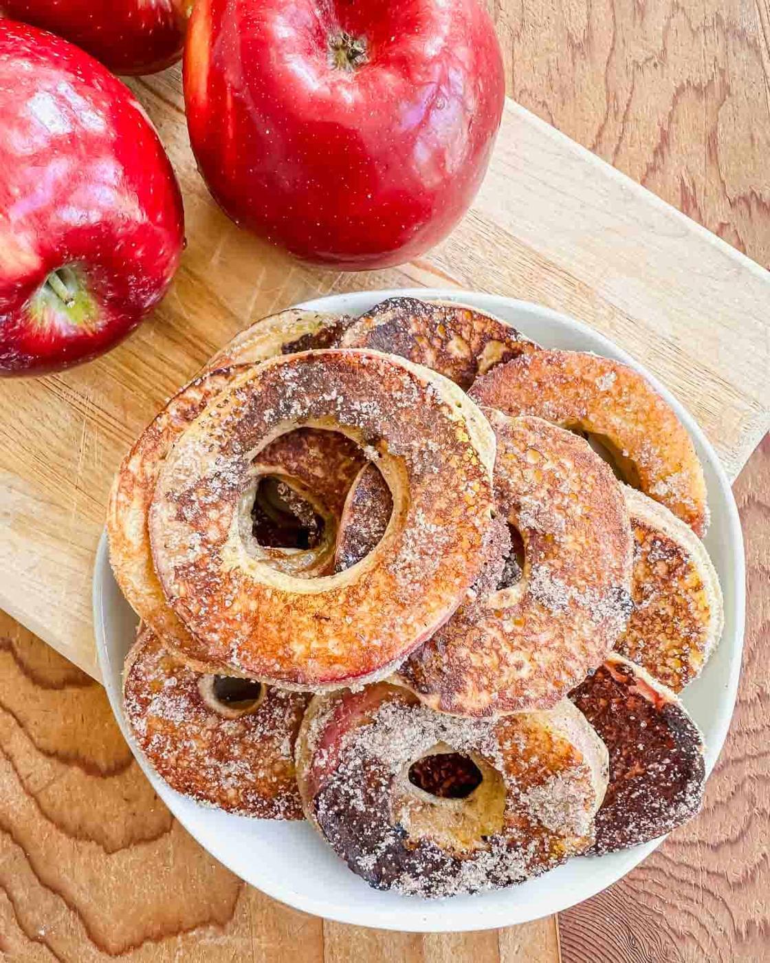 A plate of sugar-coated apple ring fritters sits on a wooden board, with two whole red apples nearby.