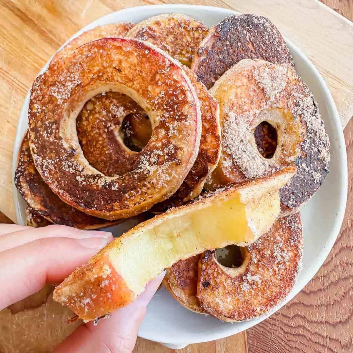 A hand holds a fried apple ring coated with cinnamon sugar above a plate stacked with more apple rings, all with holes in the center and golden brown edges.