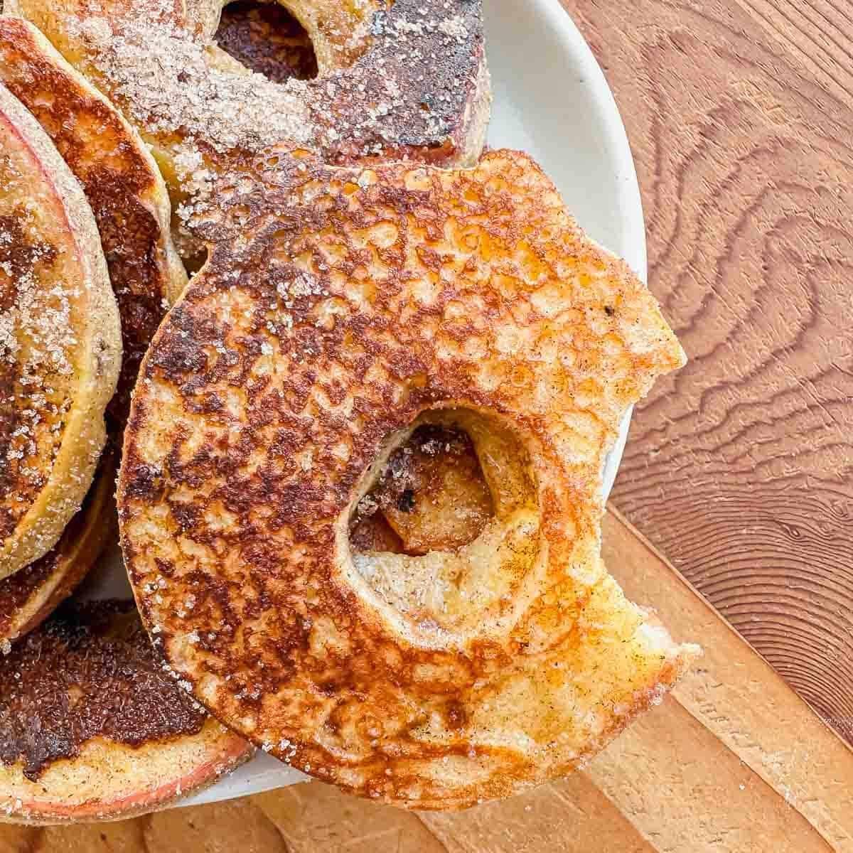 A close-up of apple slices coated in batter, fried to a golden brown, and dusted with sugar, served on a white plate atop a wooden surface. One slice has a bite taken out.