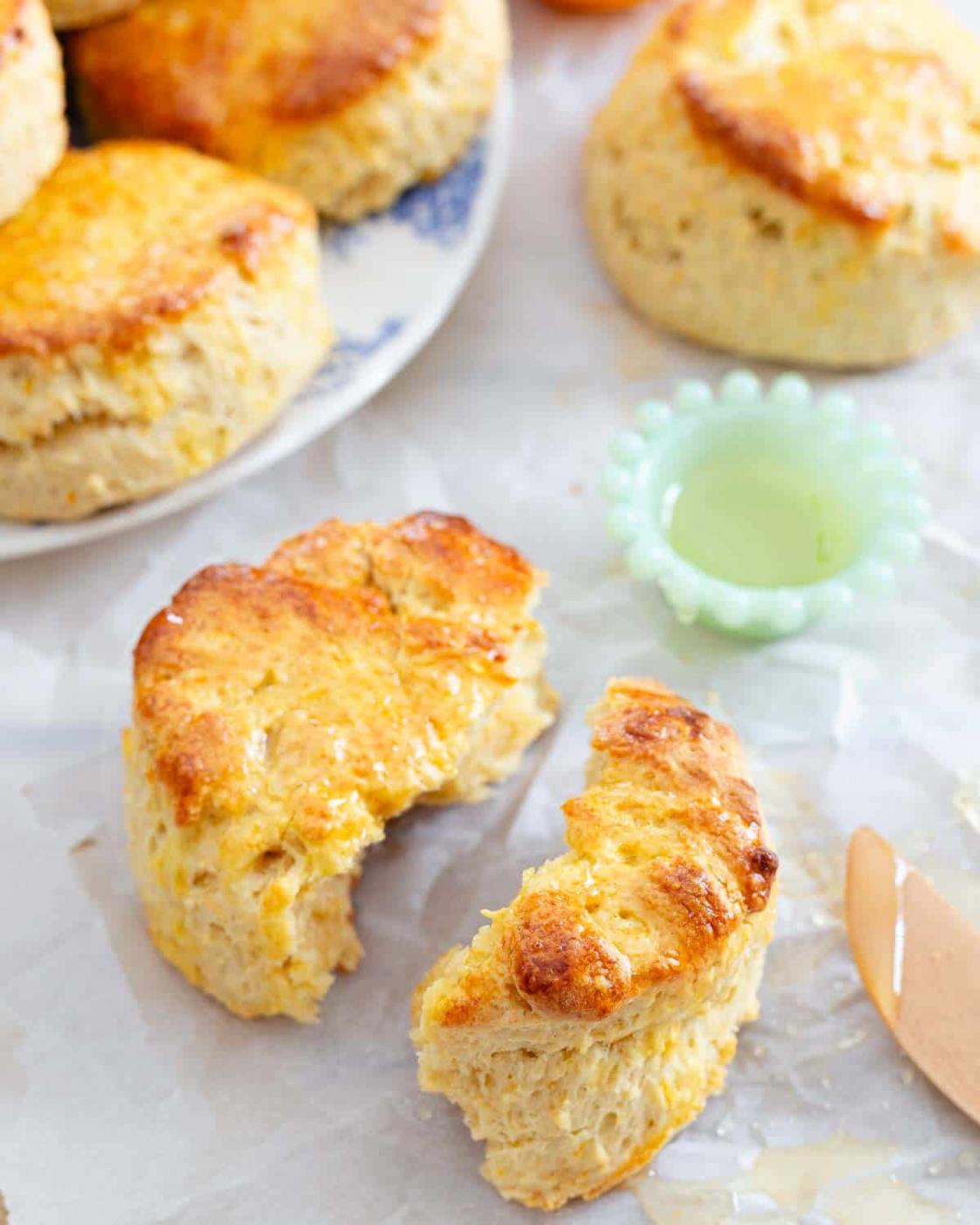 A plate with golden brown biscuits topped with honey sits in the background, with one biscuit in the foreground broken in half, revealing a fluffy interior. A small green dish and a knife are nearby.