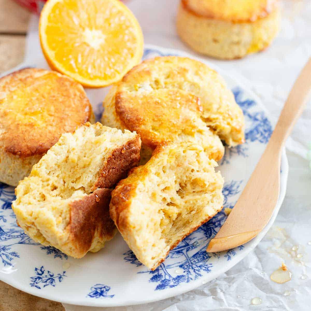 A blue and white plate with freshly baked honey biscuits, a halved orange, and a wooden knife. One biscuit is split open, revealing a fluffy texture.
