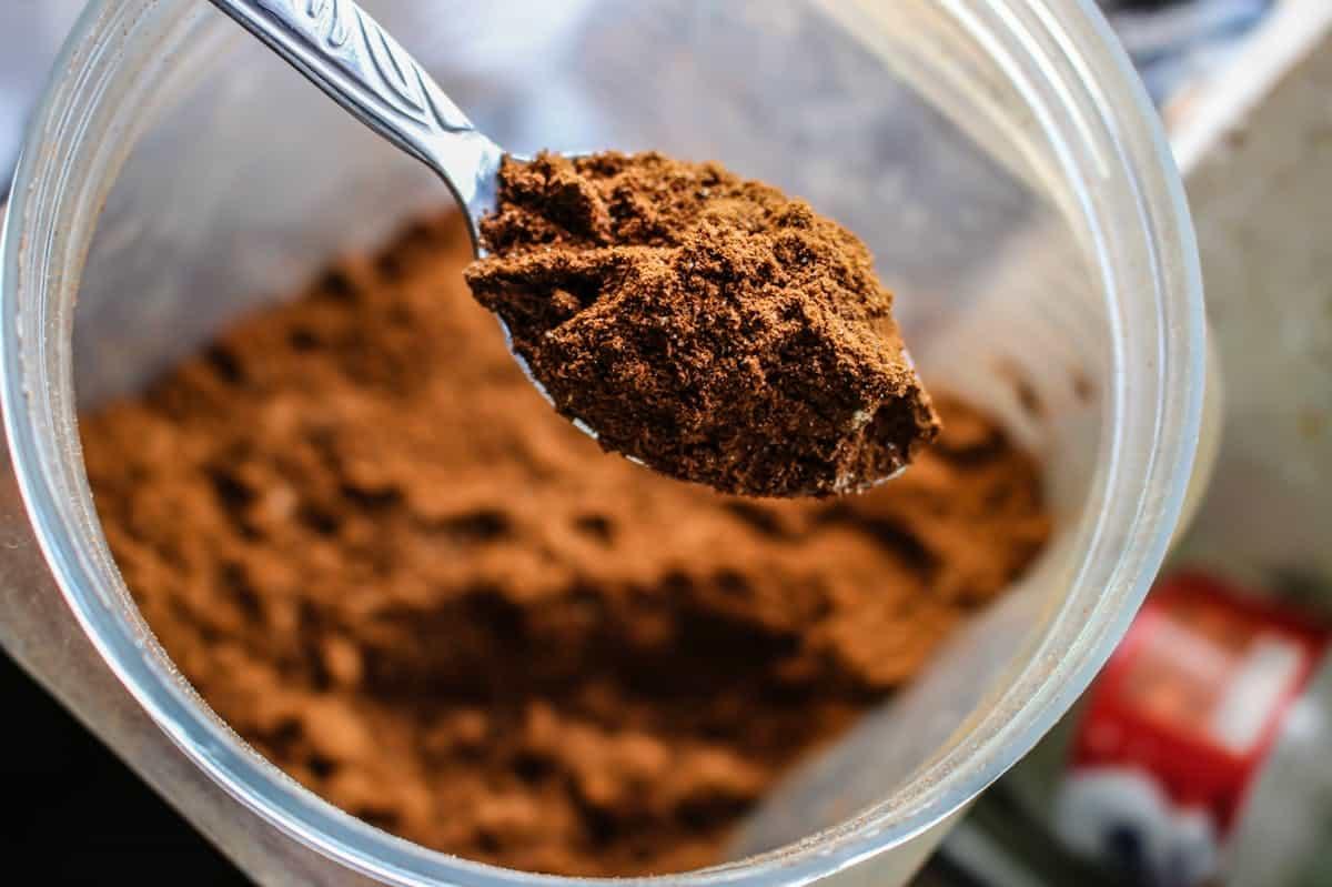 A close-up image of a spoon holding a heap of brown powder above an open jar filled with more of the same powder. The texture appears fine and loose, suggesting it might be a type of spice or powdered ingredient.