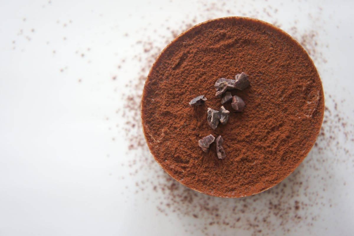 A round dessert topped with a thick layer of cocoa powder and garnished with dark chocolate chunks is placed on a white surface. Cocoa powder is lightly scattered around the dessert.