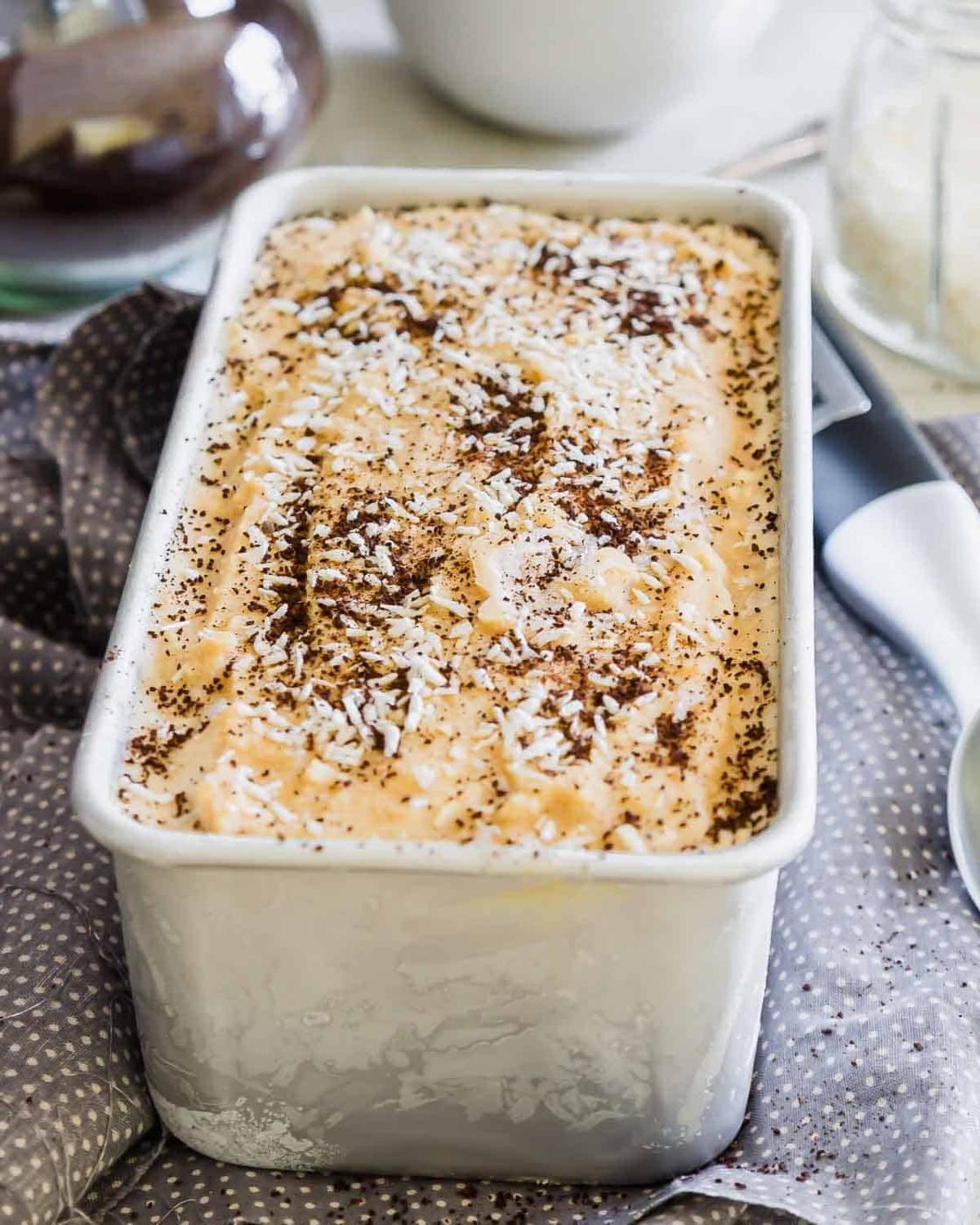 A rectangular metal container filled with creamy coffee ice cream, sprinkled with shredded coconut and coffee grinds. It sits on a gray polka dot cloth. A white scoop and a jar are partially visible in the background.
