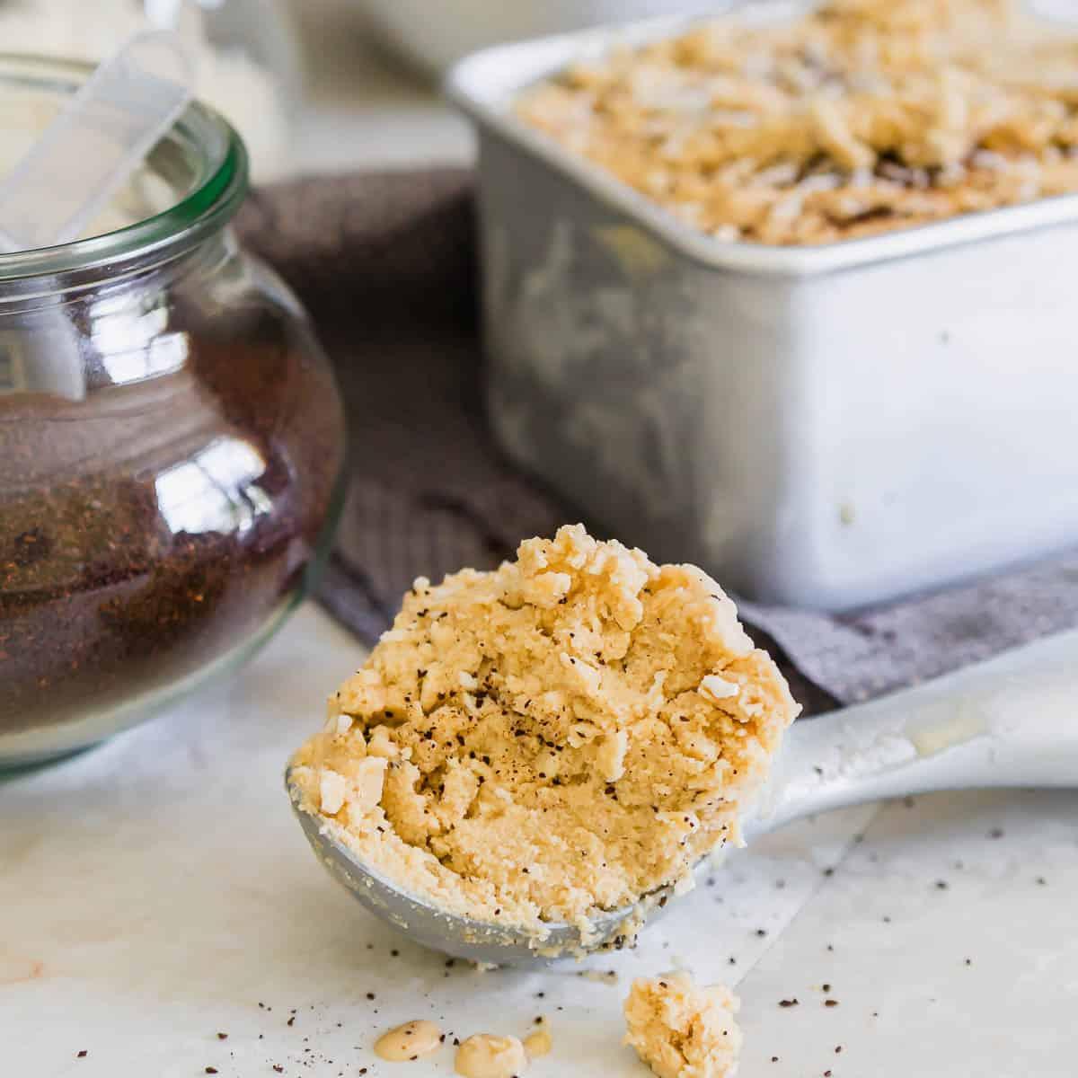 A scoop of creamy coffee ice cream sprinkled with fine coffee grounds sits on a marble surface. A metal container filled with more ice cream and a glass jar of coffee are in the background.