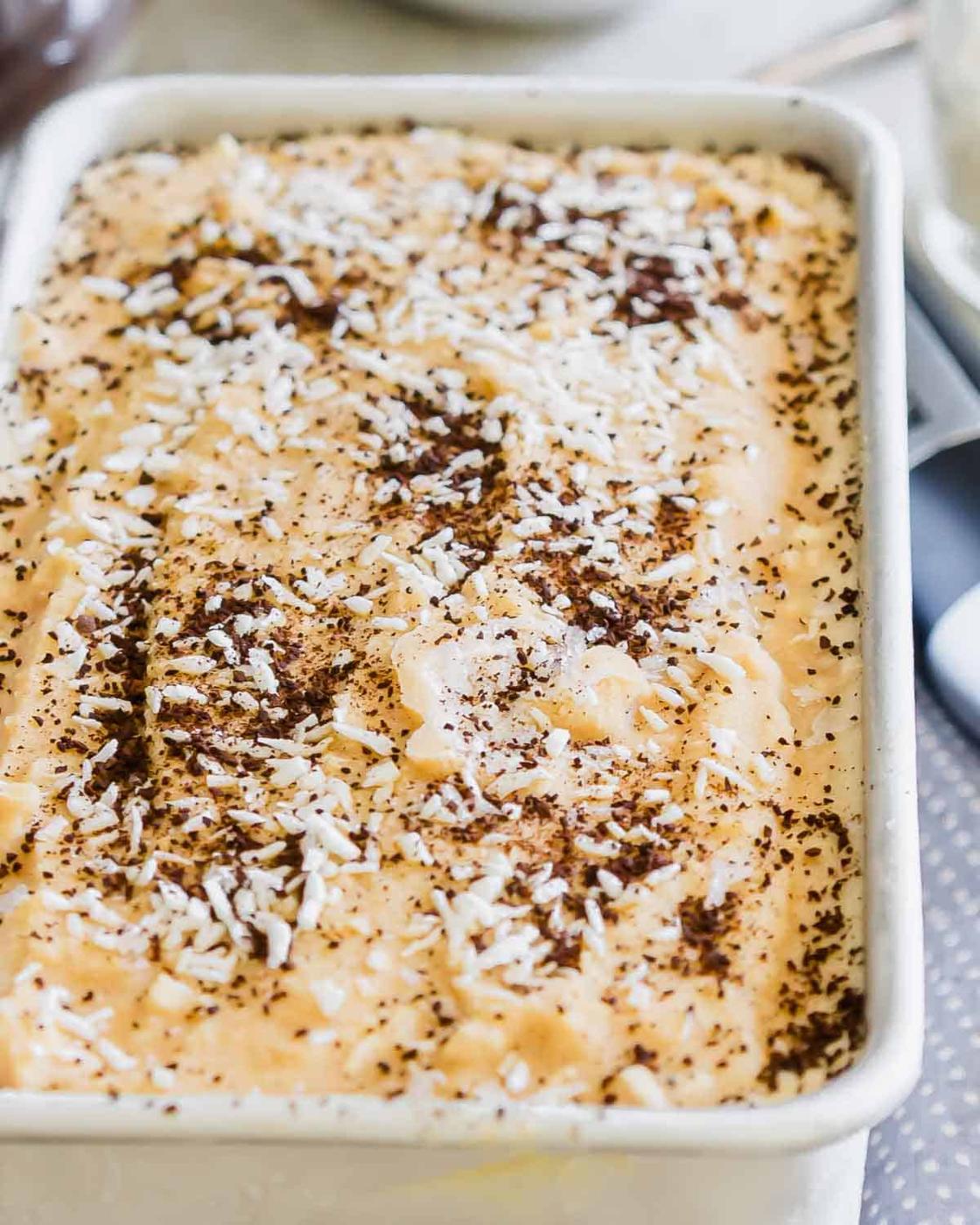 A close-up of creamy coffee ice cream with coffee grounds and white coconut flakes, served in a rectangular dish. The dessert has a light texture with visible swirls and sprinkled toppings for added texture.