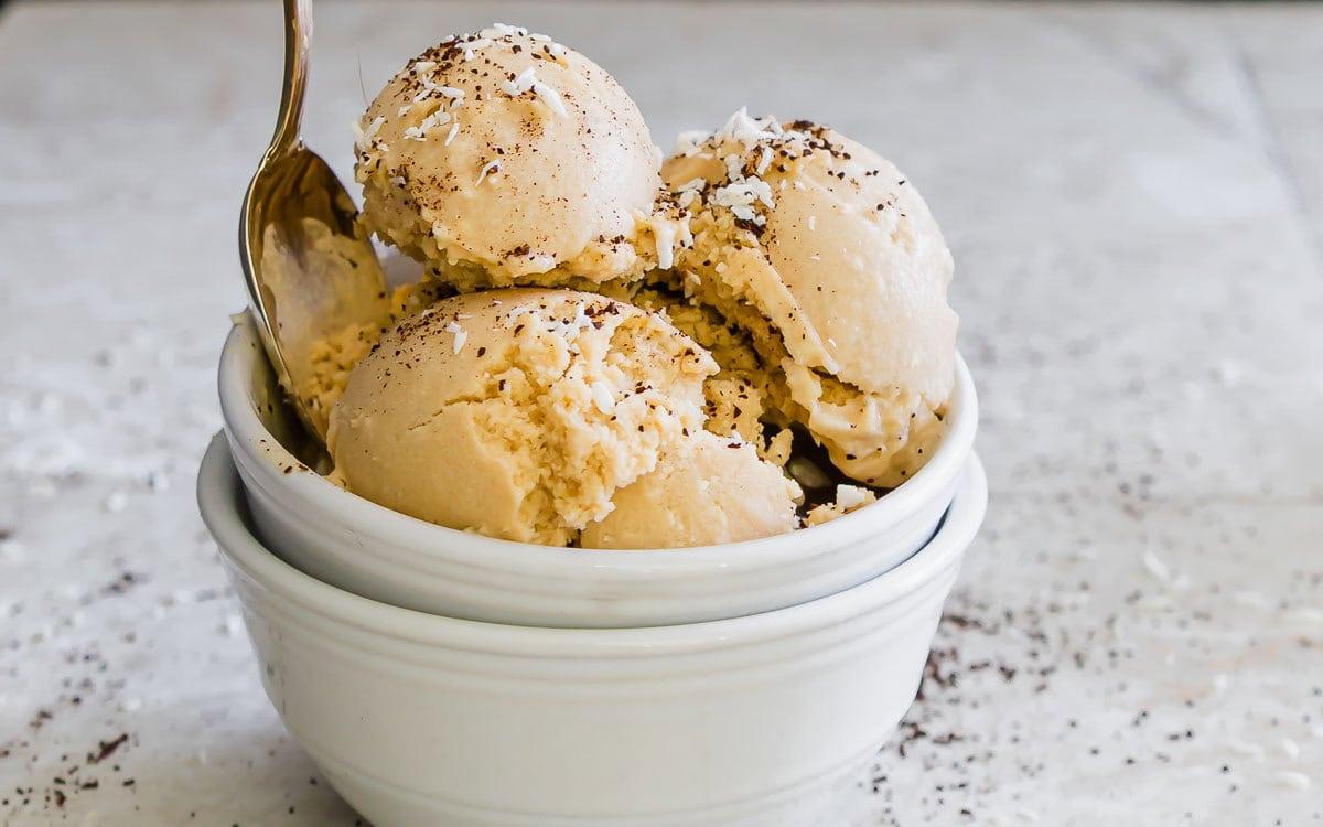 A bowl with three scoops of creamy coffee ice cream, sprinkled with coffee grinds and small coconut pieces. A spoon is resting in the bowl. The bowl is placed on a speckled surface.