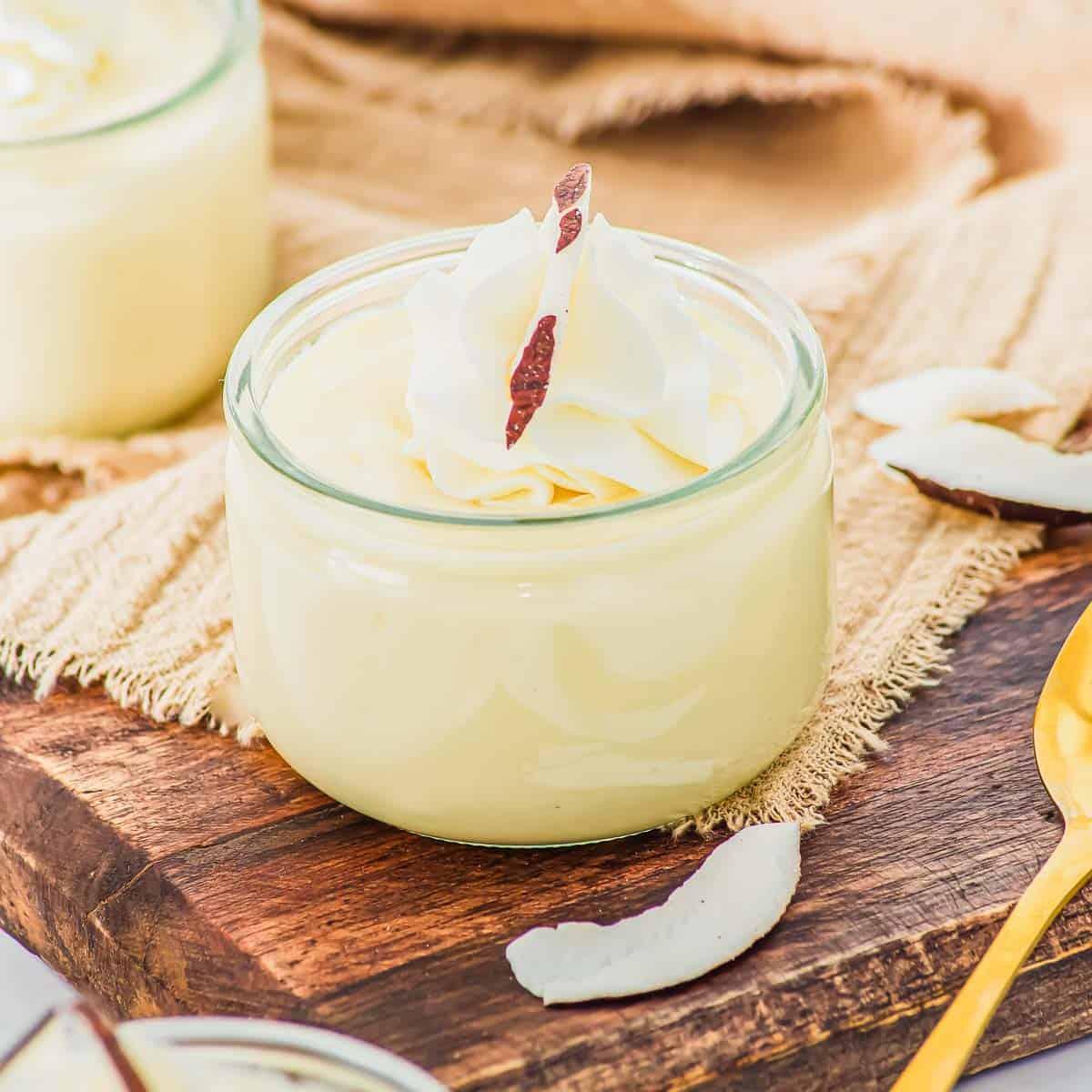 A small glass jar filled with coconut pudding, topped with whipped cream and a coconut shard, sits on a wooden board next to coconut flakes and a gold spoon.