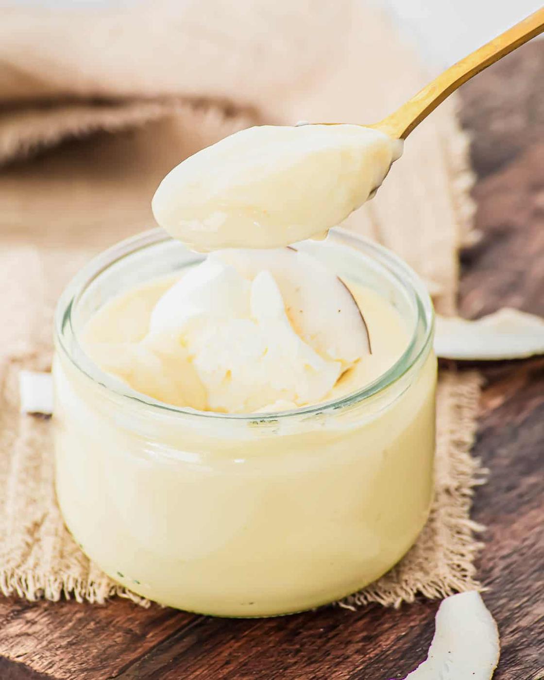 A spoon holding creamy, pale yellow coconut pudding hovers above an open glass jar filled with more custard, set on a piece of burlap atop a wooden surface. Coconut flakes are scattered nearby.