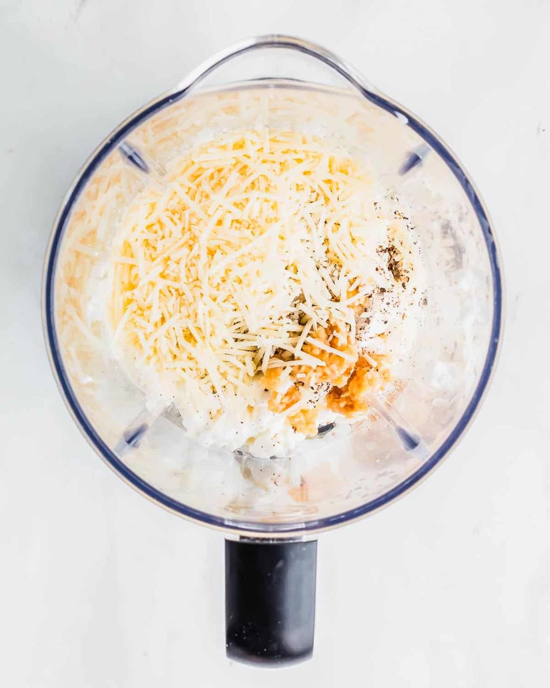 Top view of a blender with ingredients including shredded cheese, cottage cheese, and seasonings inside. The blender is placed on a light surface.