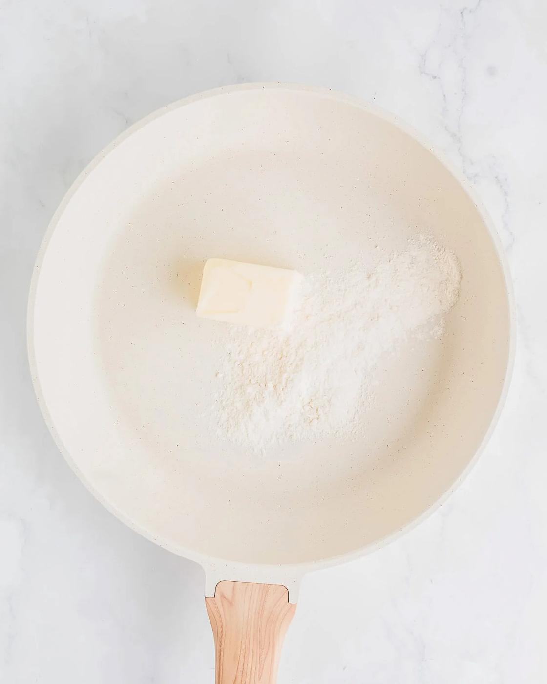 A white frying pan on a marble surface contains a small pile of flour and a pat of butter, ready for cooking. The pan has a wooden handle.