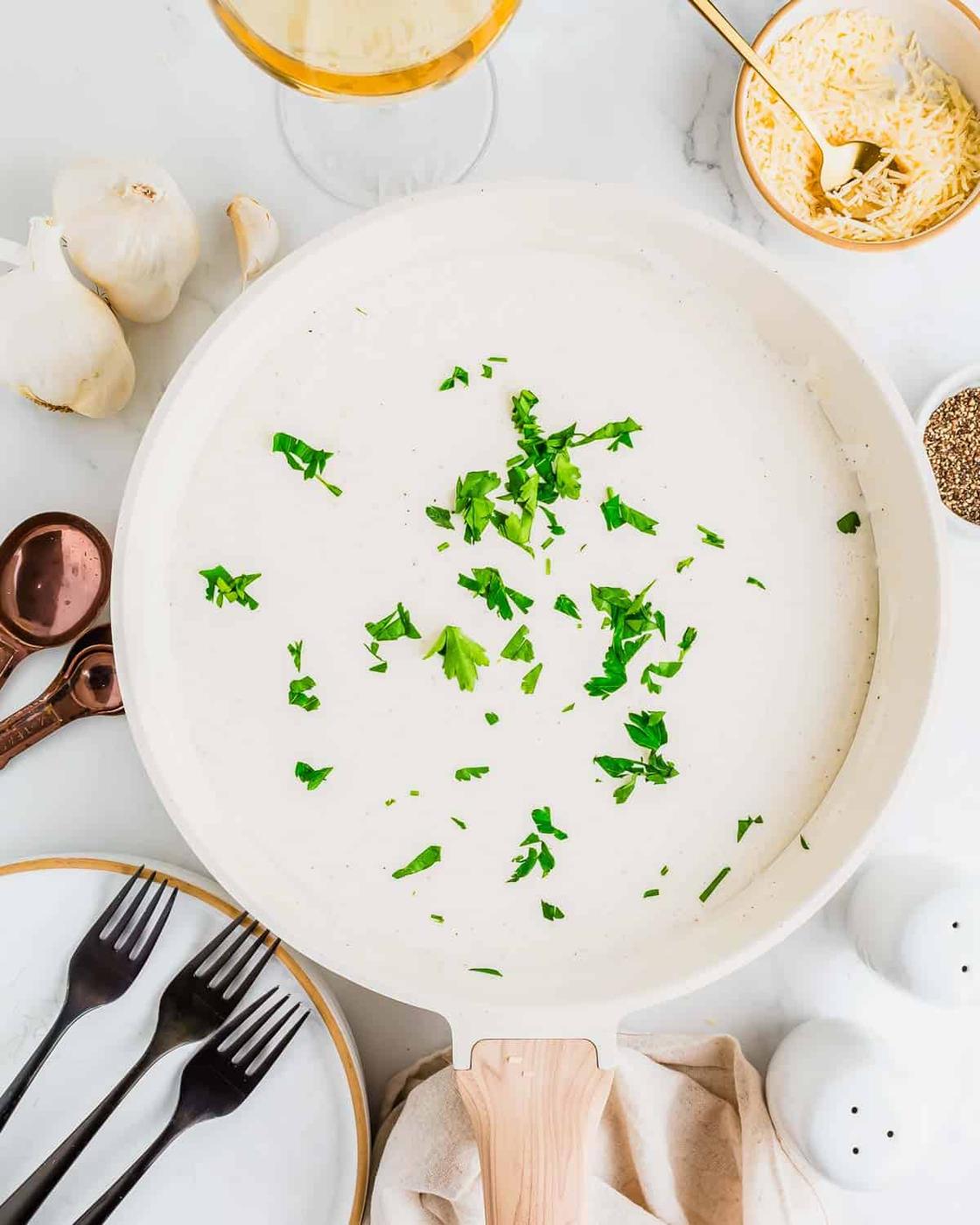 A white skillet filled with thickened cottage cheese Alfredo sauce, topped with chopped parsley. Surrounding the skillet are garlic cloves, a bowl of grated cheese, black pepper, copper measuring spoons, a plate with forks, and a glass with a golden drink.