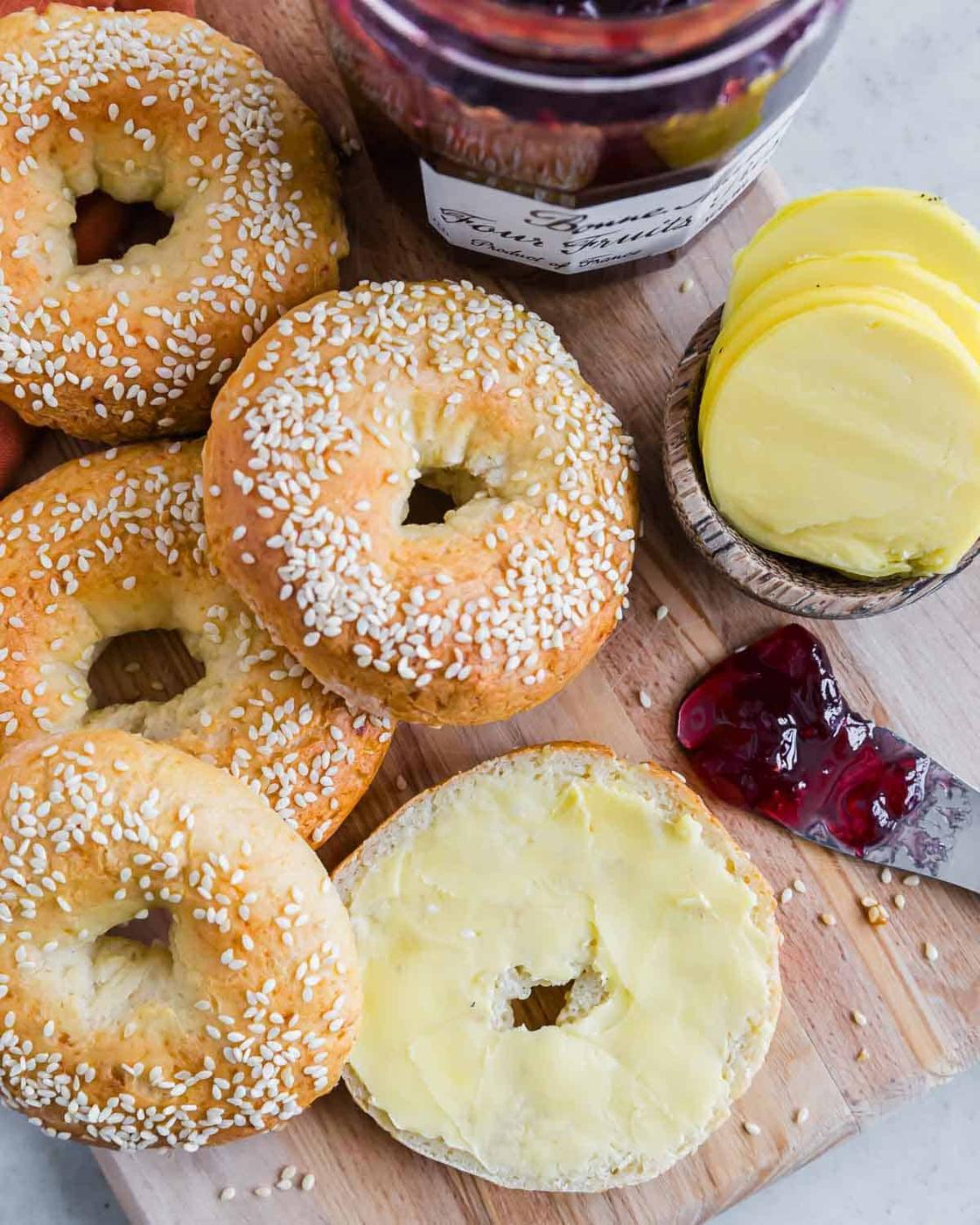 A wooden board holds several sesame-seed cottage cheese bagels, a sliced bagel with butter, a jar of berry jam, and slices of yellow cheese in a small bowl. A dollop of jam is spilled next to the bagels.