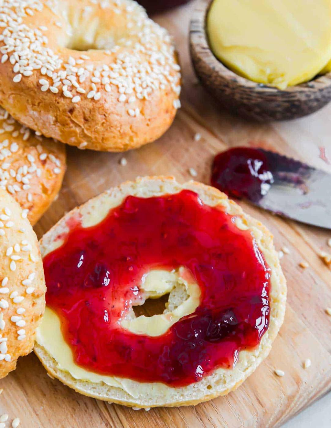 A sliced bagel topped with butter and berry jam sits on a wooden board. Several whole sesame bagels are in the background. A knife with jam and a wooden bowl with butter are to the side.