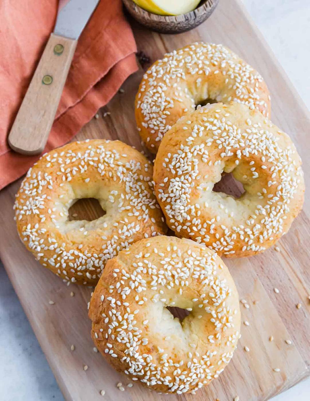 Four sesame seed-covered gluten-free protein bagels are placed on a wooden cutting board with an orange napkin and a knife beside them.